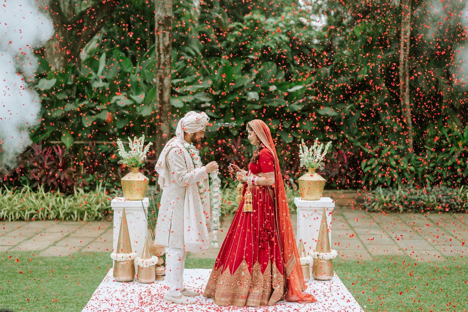 Indian bride in red lehenga and groom in white sherwani exchange vows at outdoor wedding ceremony with floral decor.