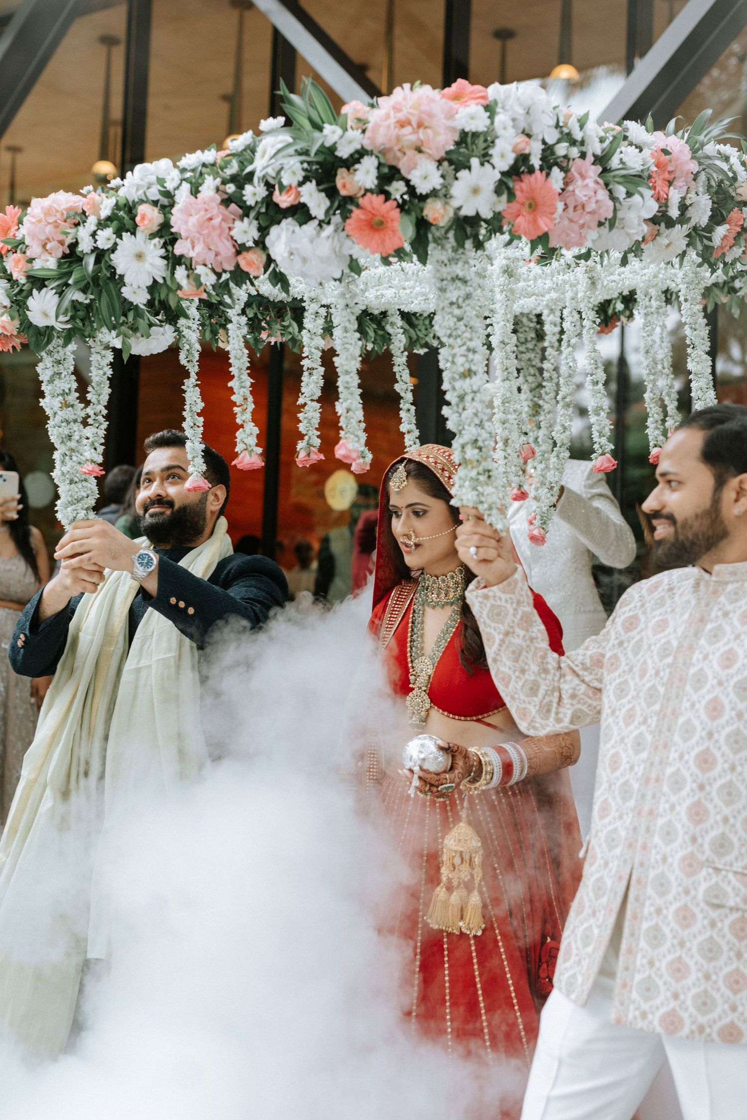 Indian bride in red lehenga walks under floral phoolon ki chadar at traditional Hindu wedding ceremony.