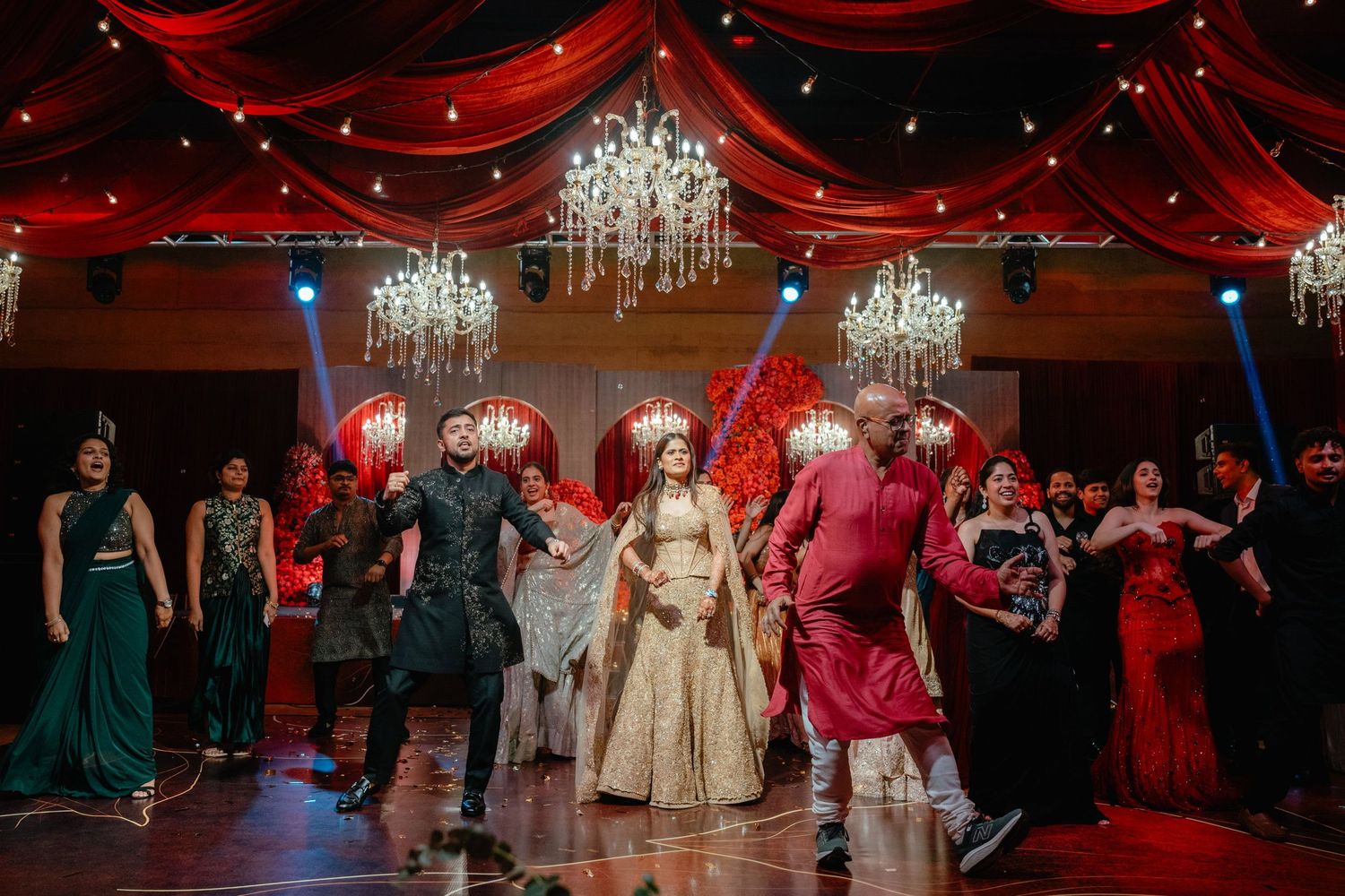 Indian wedding reception dance floor with bride in gold lehenga surrounded by guests under crystal chandeliers and red draping.
