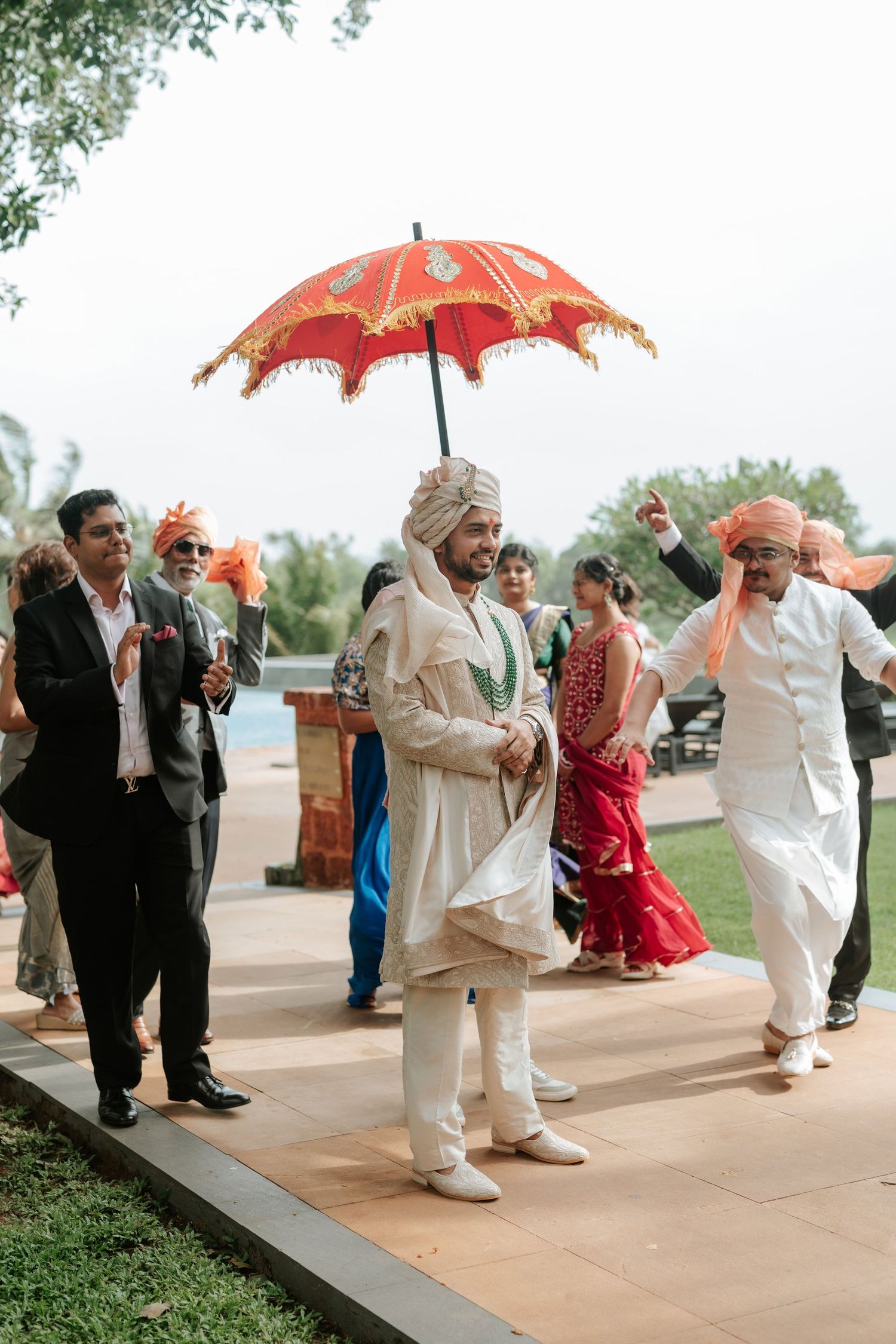 Indian groom in white sherwani walks under red ceremonial umbrella during traditional baraat wedding procession.