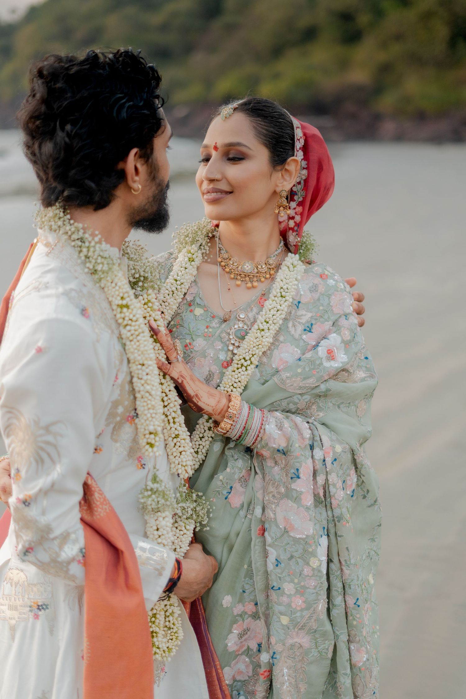 Indian couple in traditional wedding attire exchange garlands on a beach, bride in floral saree with gold jewelry.