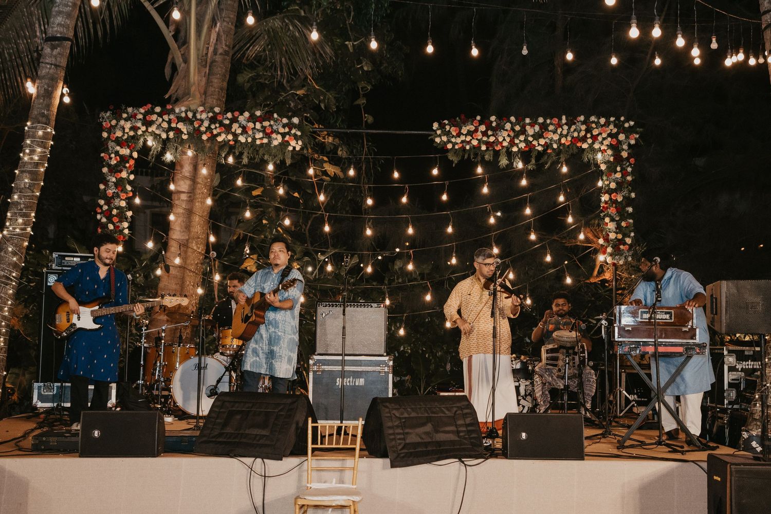 Live band performing on an outdoor stage decorated with string lights and floral backdrop at a night event.