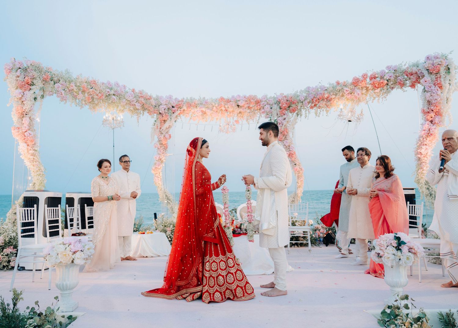A couple celebrates their traditional Indian wedding ceremony under a floral canopy by the sea during sunset.
