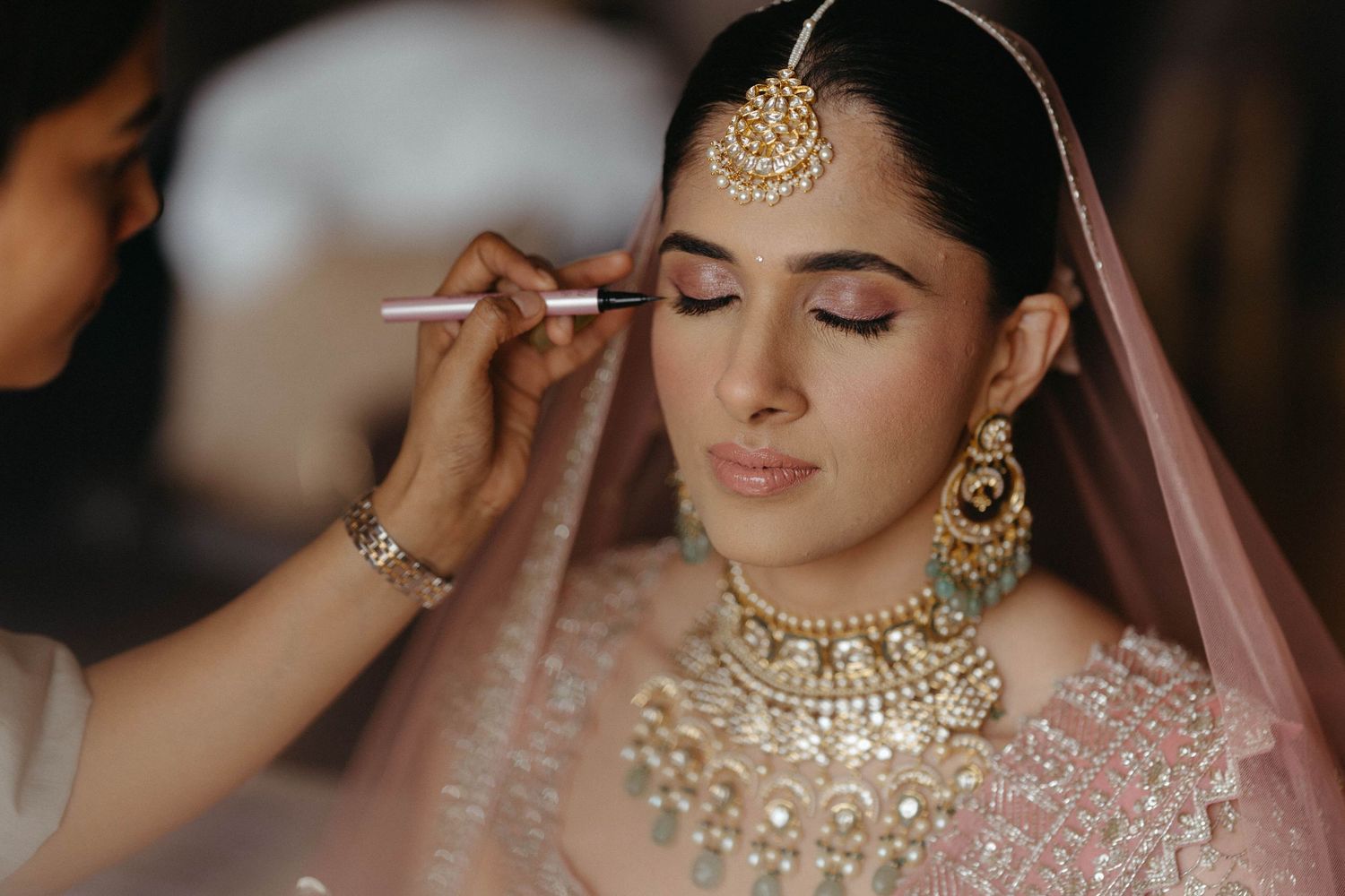 A bride wearing traditional Indian bridal jewelry and pink attire gets her makeup done during wedding preparations.