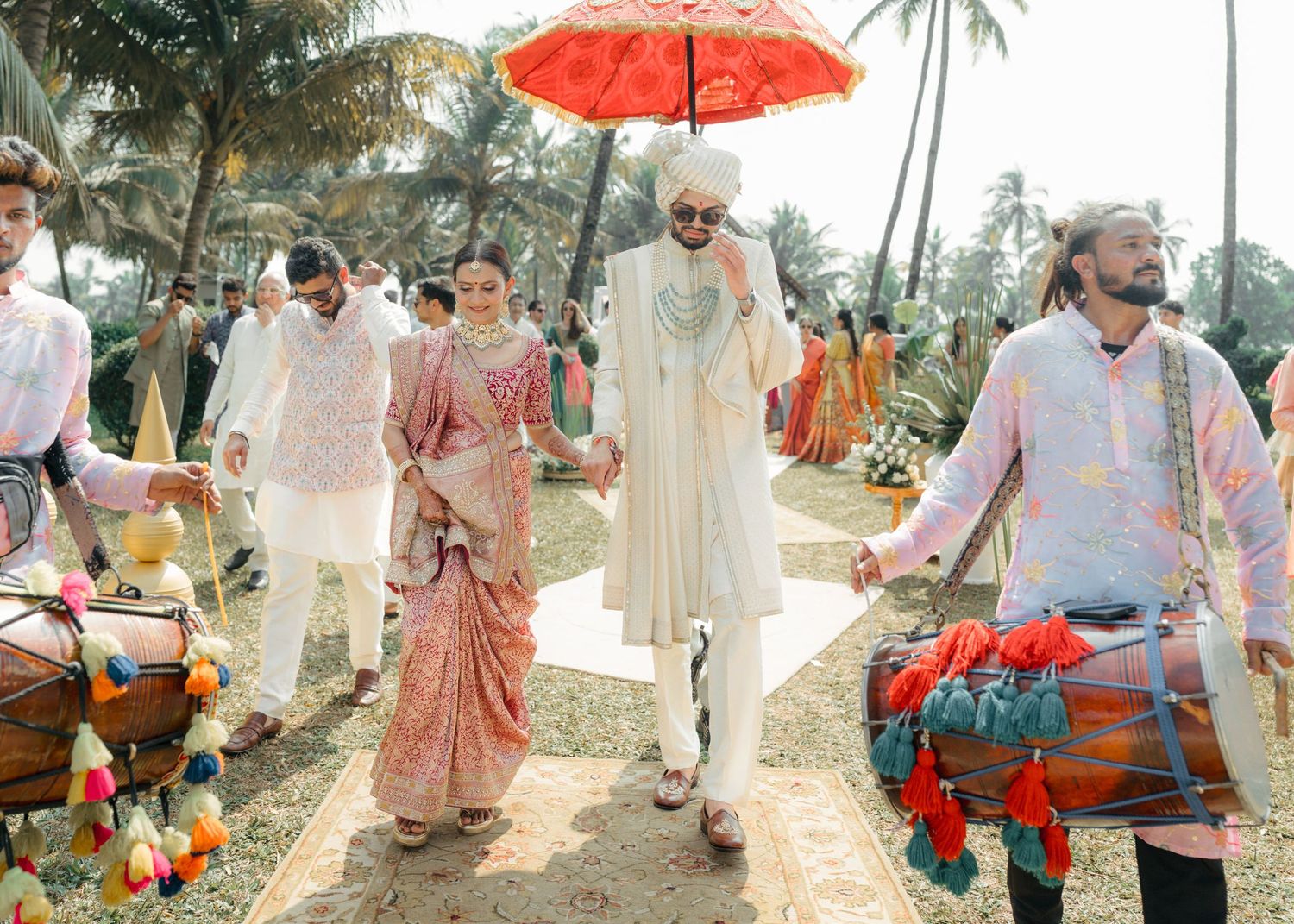 A festive outdoor procession with people in traditional Indian attire walking under a red umbrella amidst palm trees.