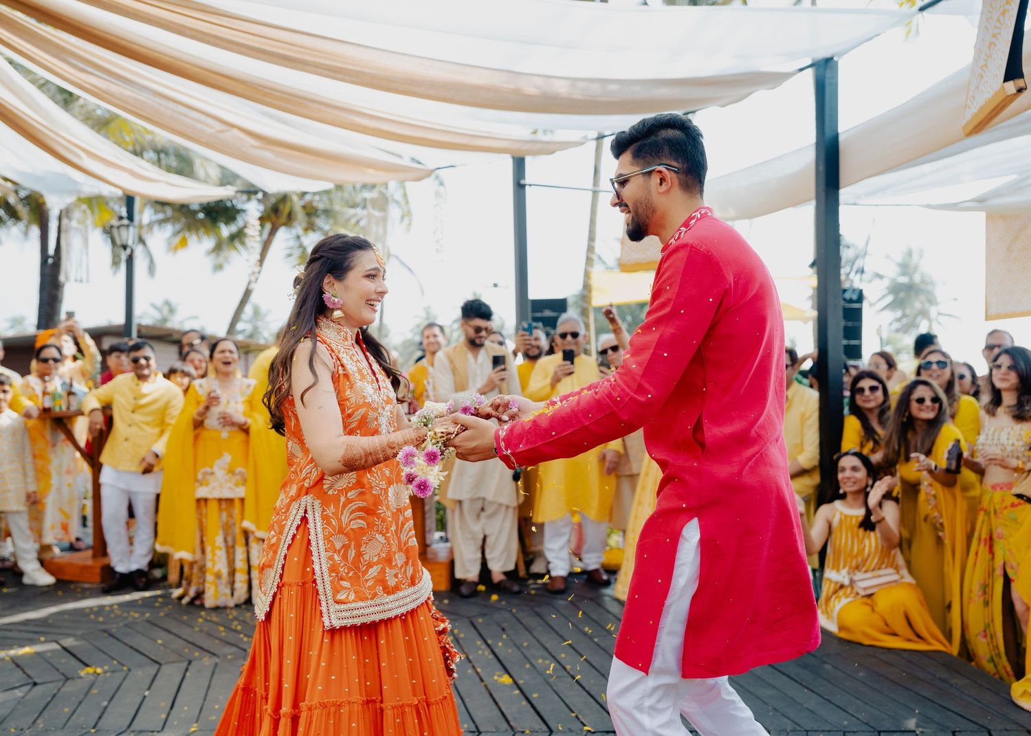 Two people dance together at a traditional Indian wedding celebration surrounded by guests wearing yellow outfits.