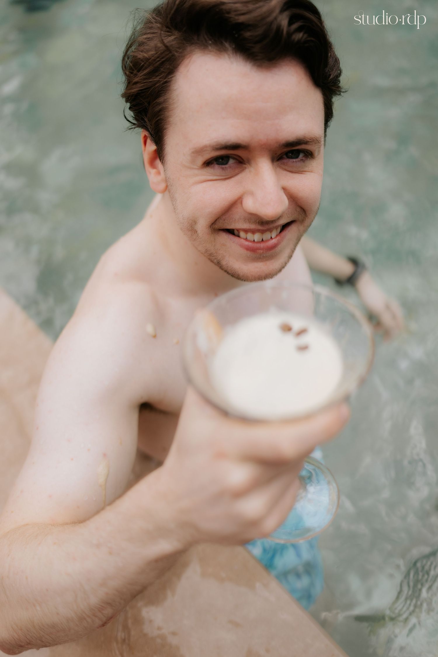 Smiling young man in a pool holding a creamy cocktail drink toward the camera in a cheerful outdoor setting.