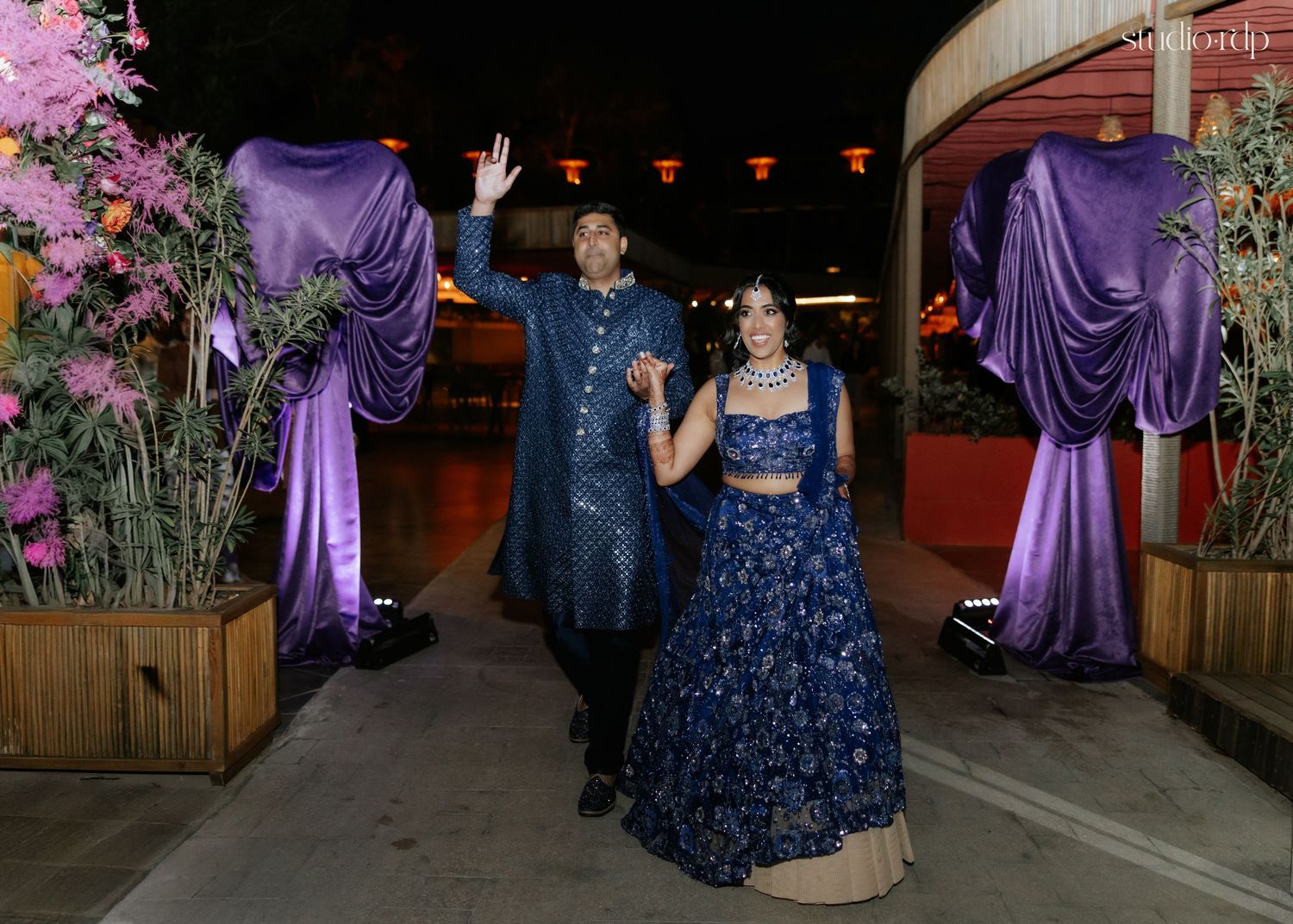 Couple in matching blue embroidered Indian outfits walking through a floral entrance with purple drapes at night.