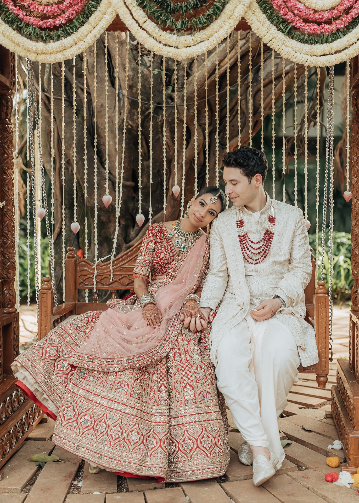 A couple in traditional Indian wedding attire sits beneath ornate hanging decorations and string lights.