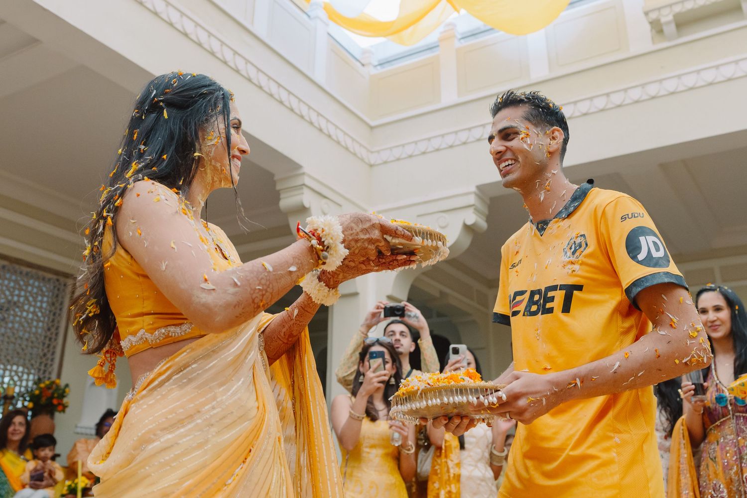 A couple in yellow attire participates in a joyful Haldi ceremony indoors.