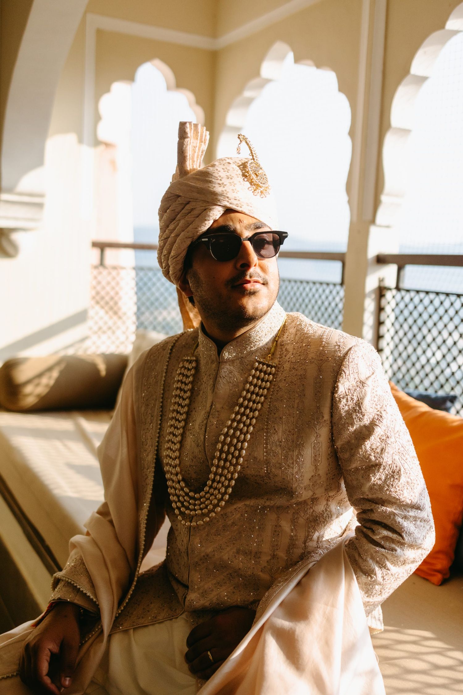 Traditional Indian groom wearing an ornate cream sherwani and turban sits in an elegant indoor setting.