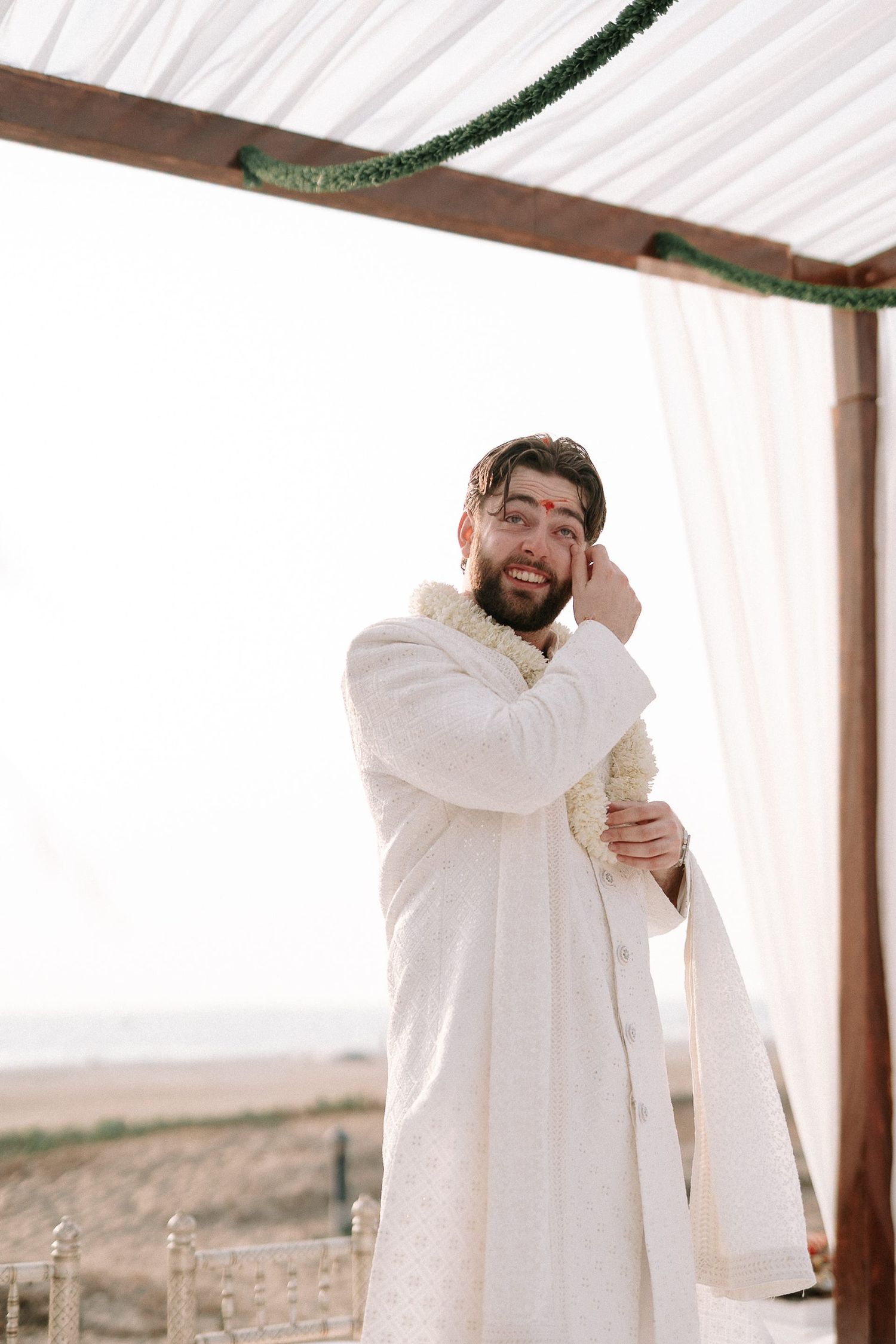 Smiling groom in white sherwani standing under wooden mandap at beach wedding ceremony.