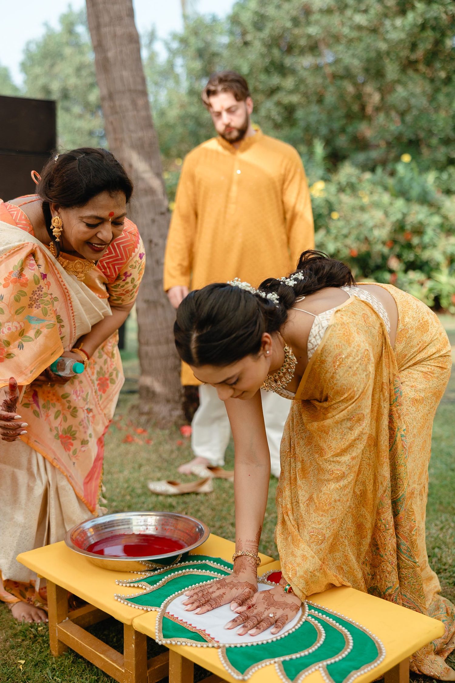 Women in colorful sarees performing traditional Indian wedding ritual with red powder and decorated table outdoors.