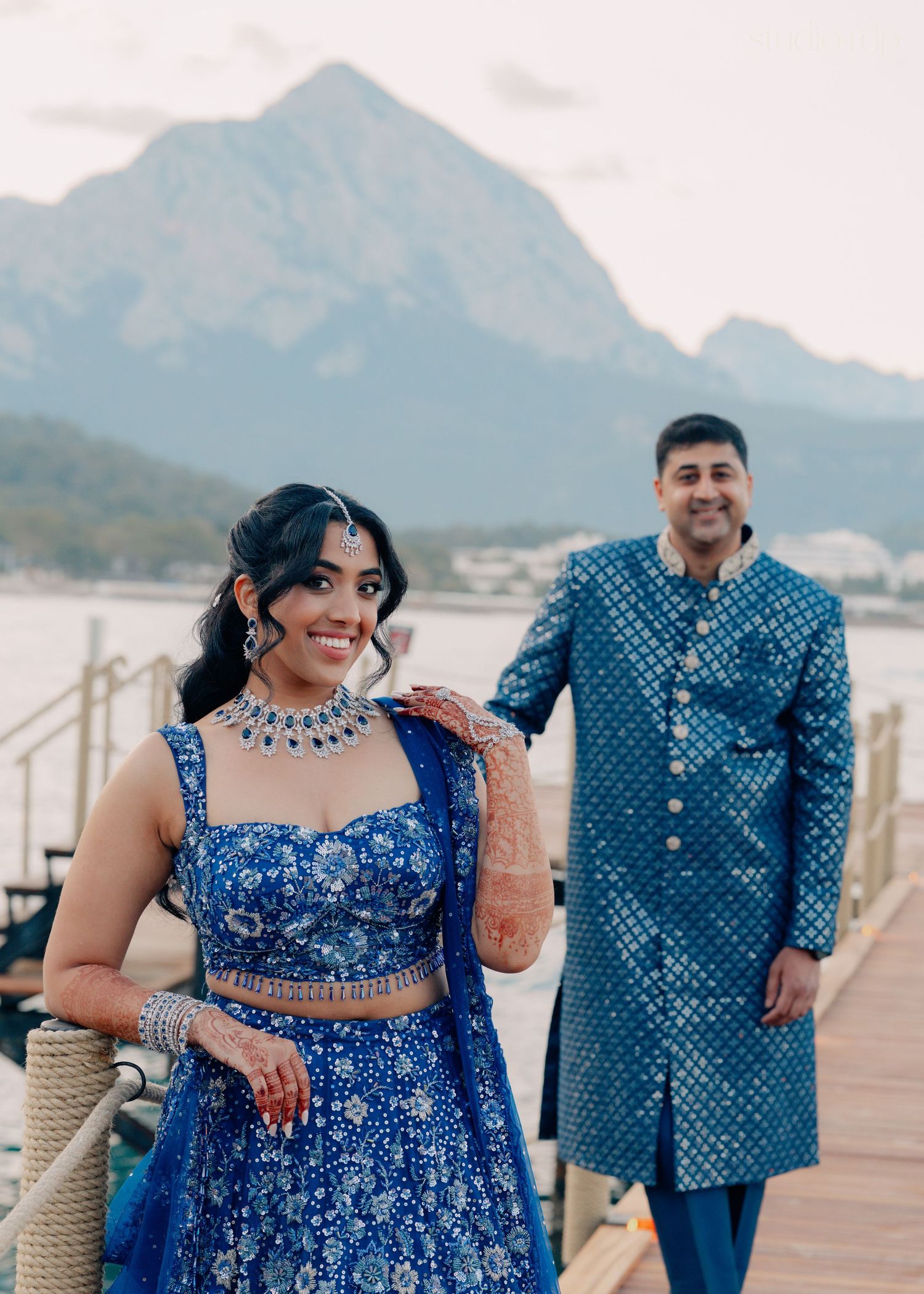 Couple in matching blue Indian wedding attire posing on a dock with mountain and water backdrop.