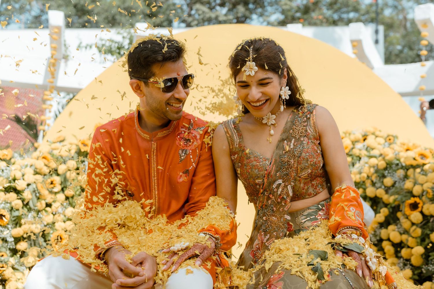 A couple in traditional Indian attire sits among marigold flowers during a wedding celebration.