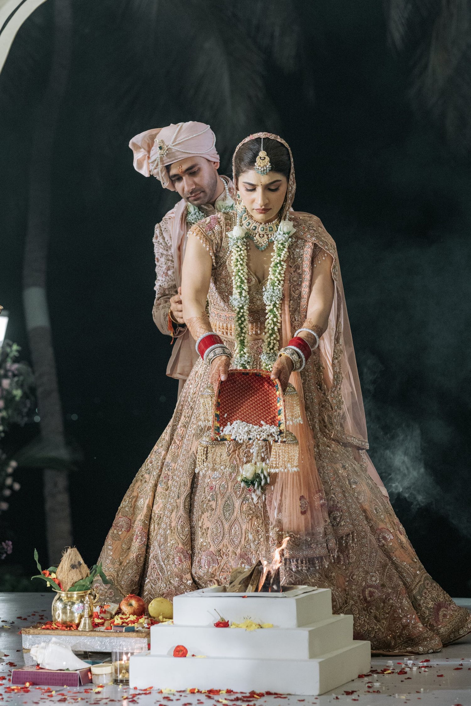 A couple in traditional Indian wedding attire performs a ritual ceremony with rose petals on a white platform.