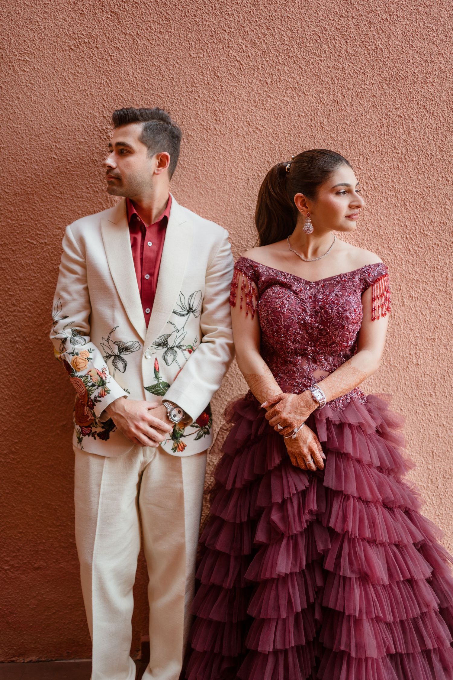 A couple in a burgundy dress and cream suit pose against a pink wall in romantic wedding attire.