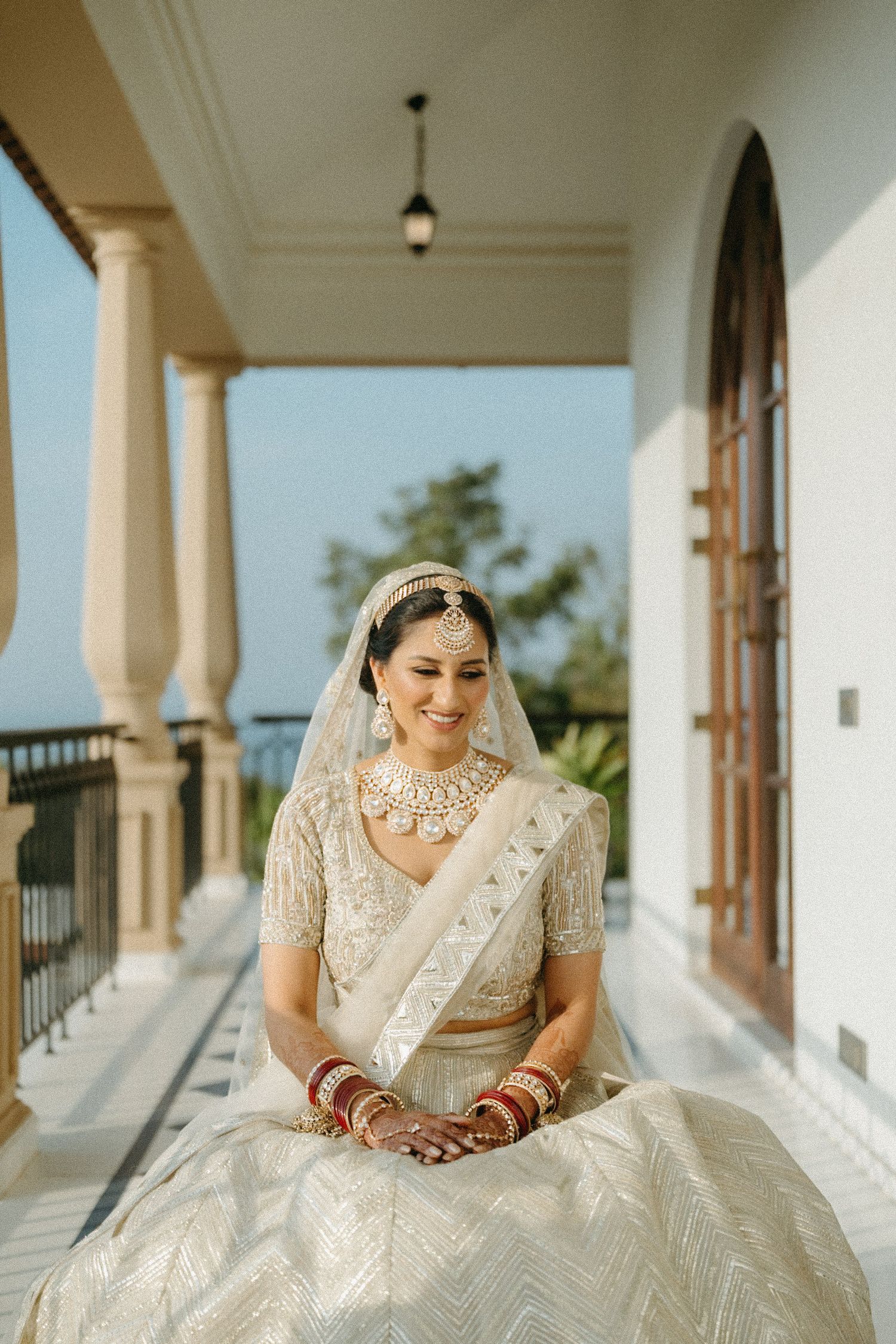 Traditional bride in white and gold lehenga poses elegantly on a covered veranda with classic architecture.