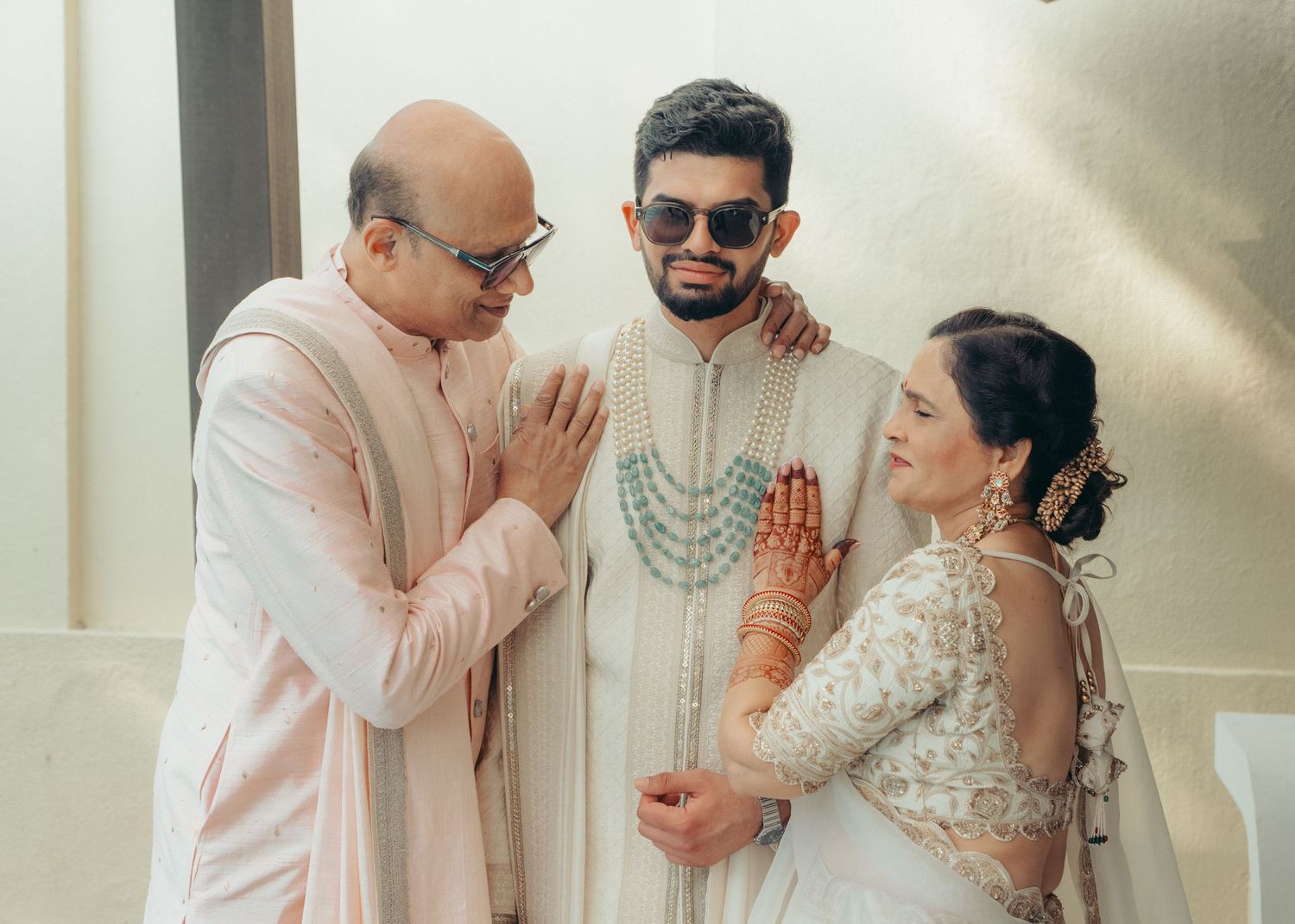 Three people in traditional Indian wedding attire pose together with greeting gestures in soft natural lighting.