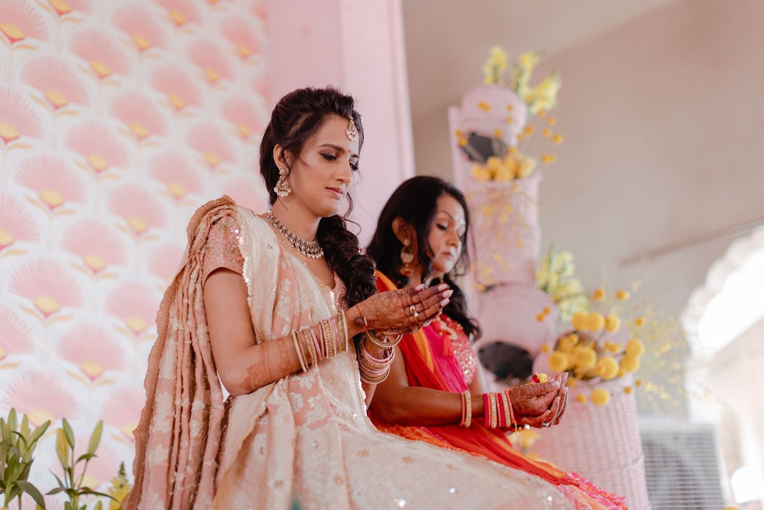 Wedding guests in traditional Indian attire check their phones during a celebration with pink decor and tiered cake.