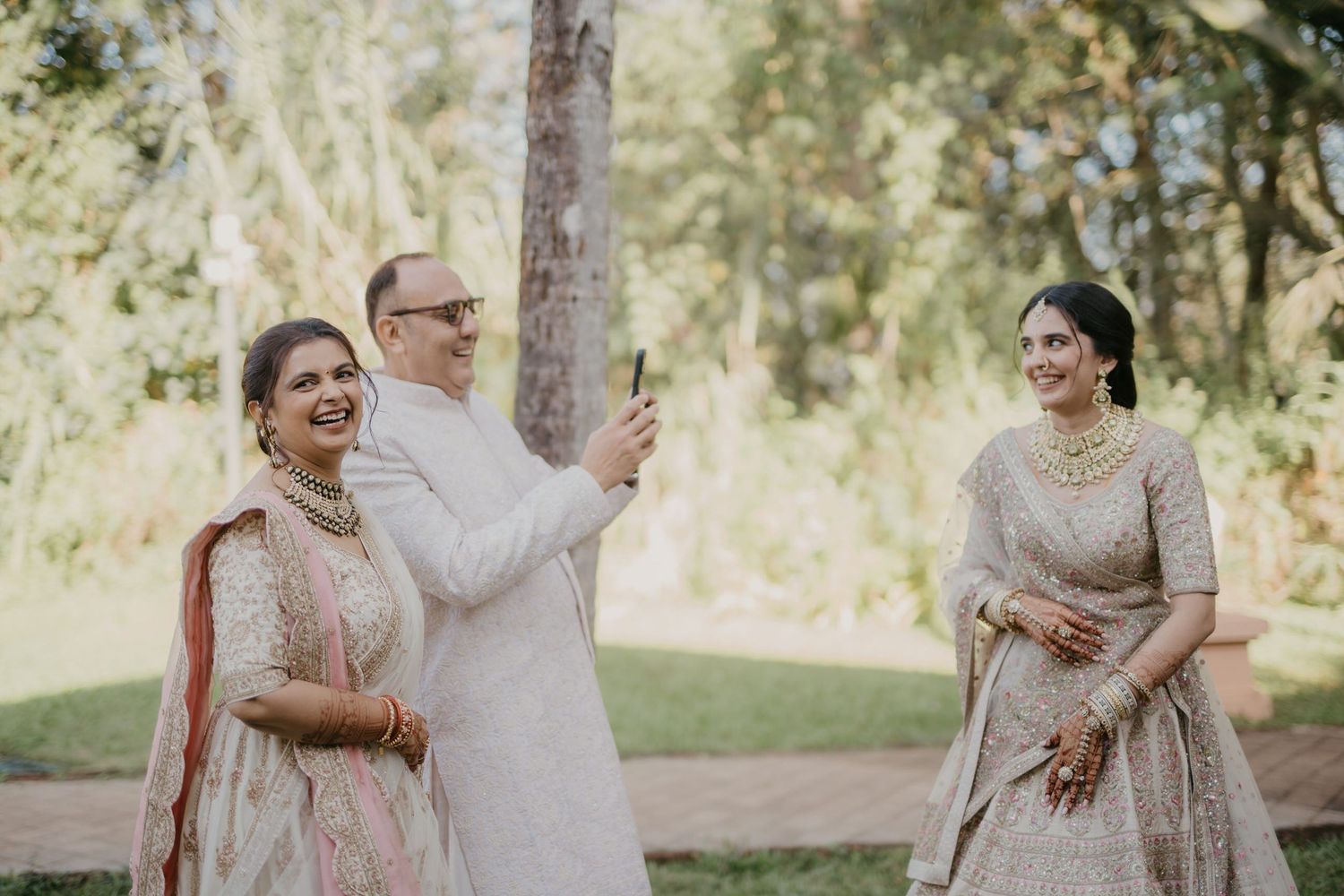 Three people in traditional South Asian wedding attire share a candid moment outdoors in a sunlit garden setting.