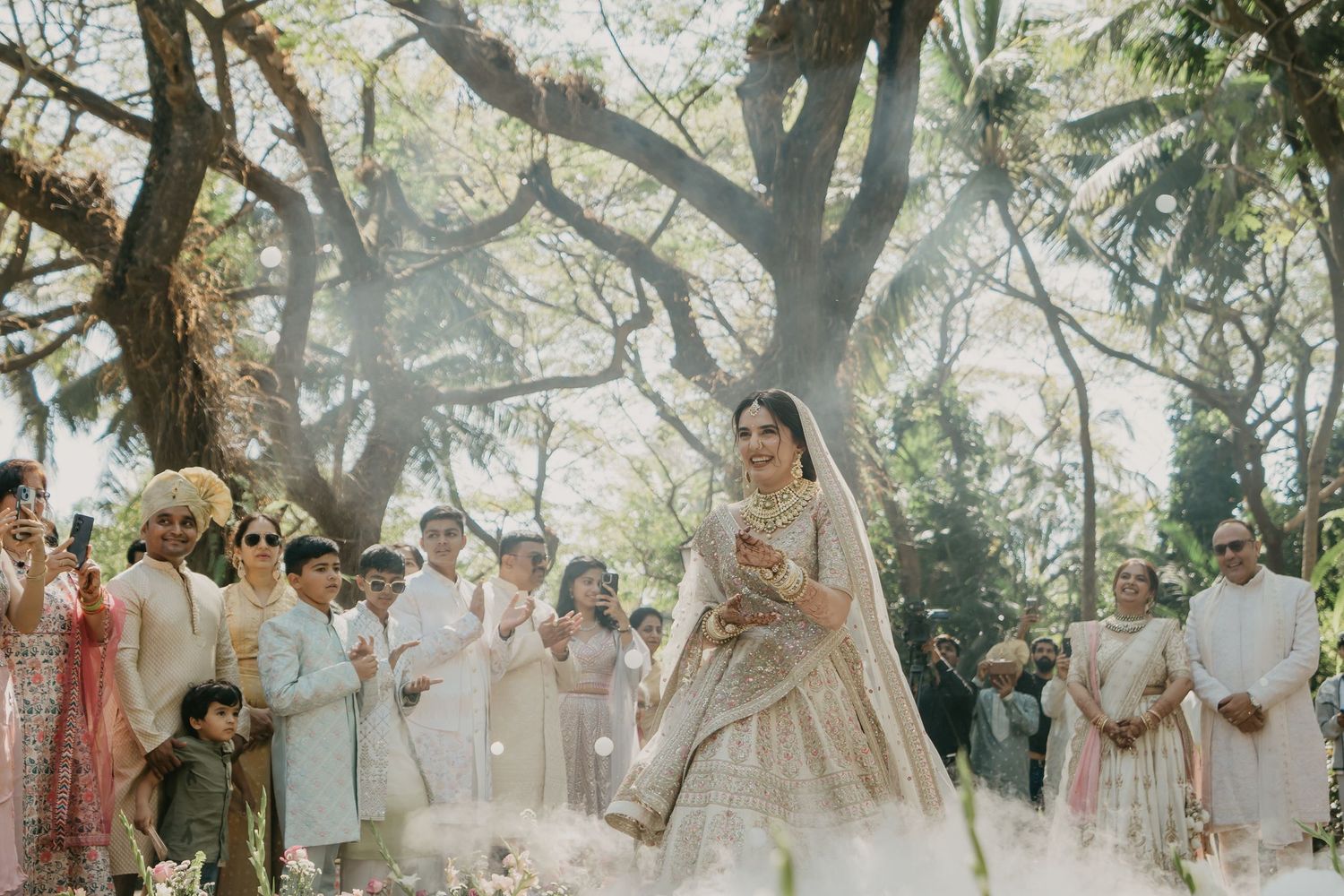 Wedding party poses together in sun-dappled outdoor garden setting with soft light filtering through trees.