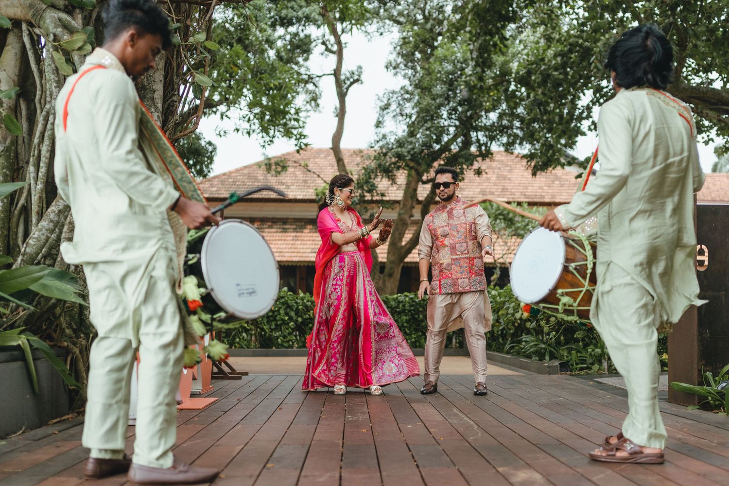 A bride in red lehenga dances on wooden deck surrounded by dhol players in traditional Indian wedding celebration.