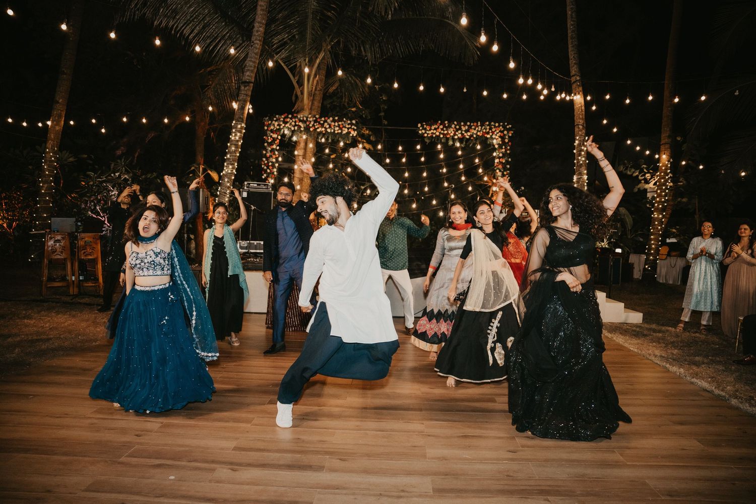 Guests dancing joyfully at an outdoor Indian wedding reception under string lights at night.