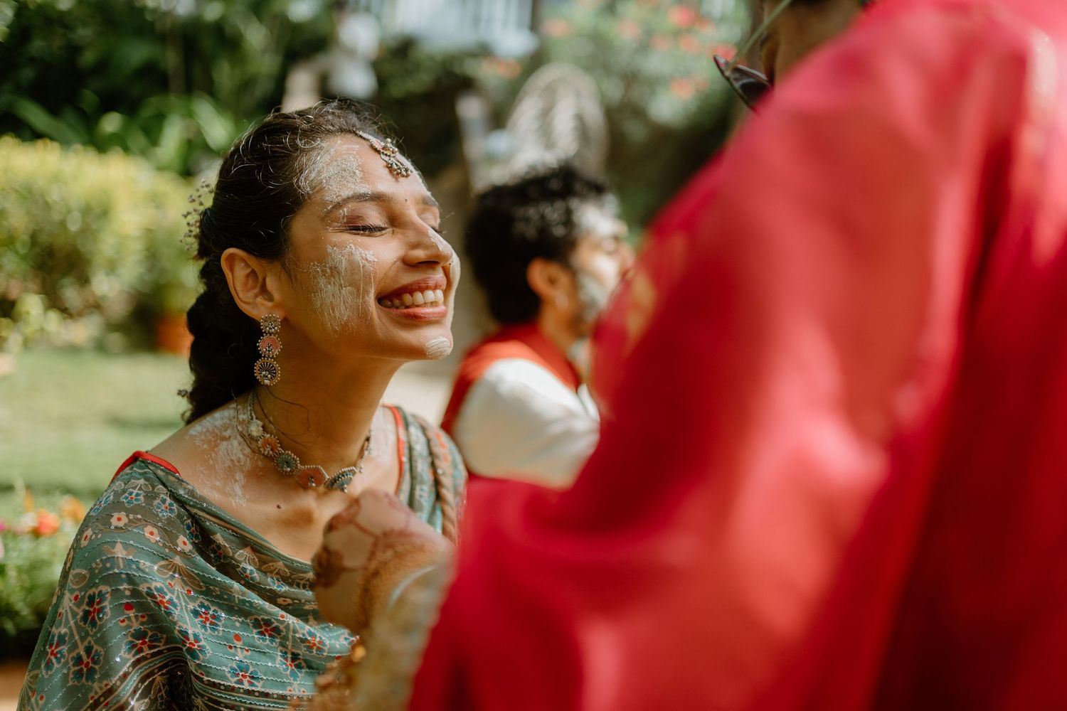 A joyful Indian bride laughs during a Haldi ceremony, her face covered in turmeric paste, wearing traditional jewelry.