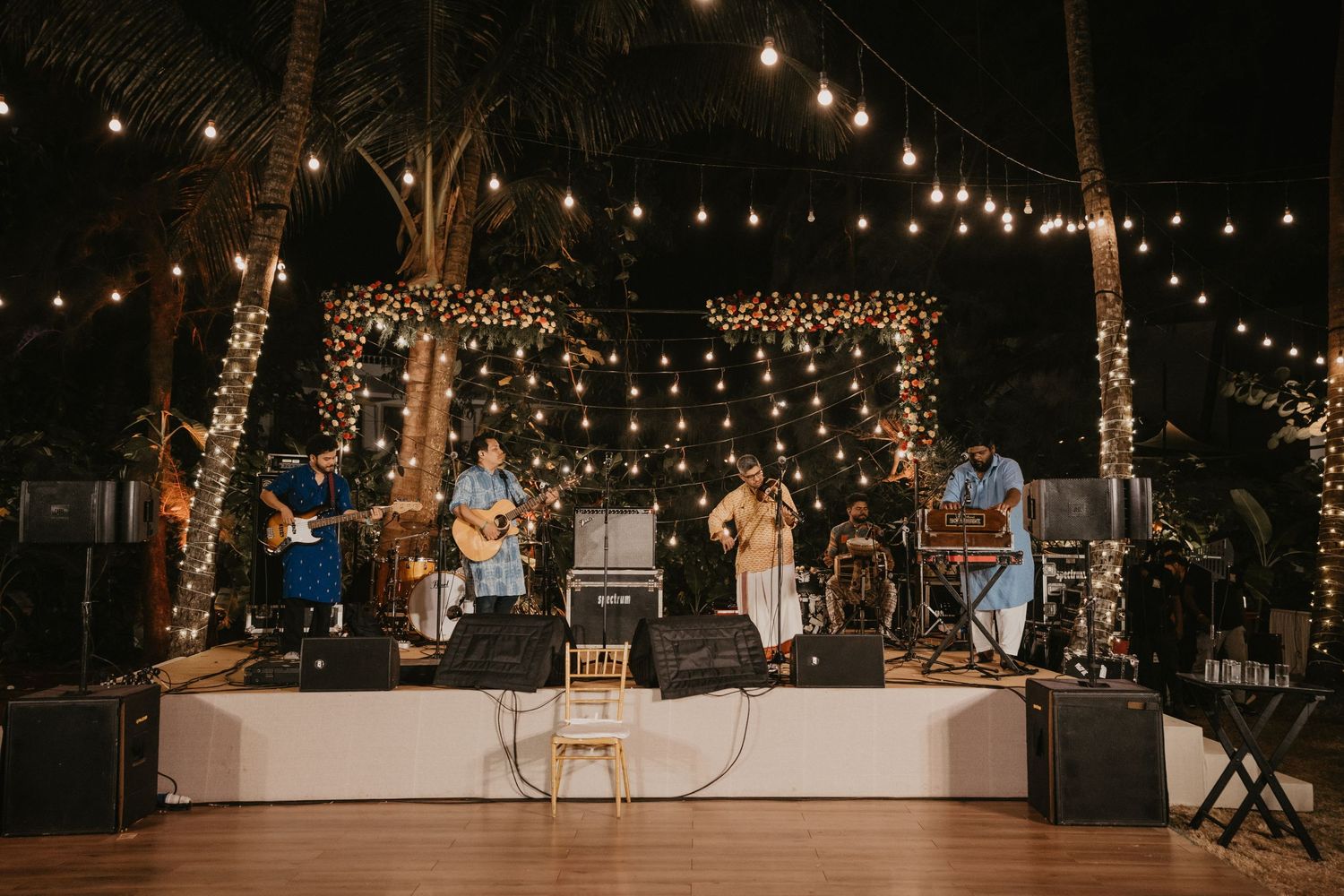 Live band performing on a decorated outdoor stage with string lights and palm trees at a tropical evening event.