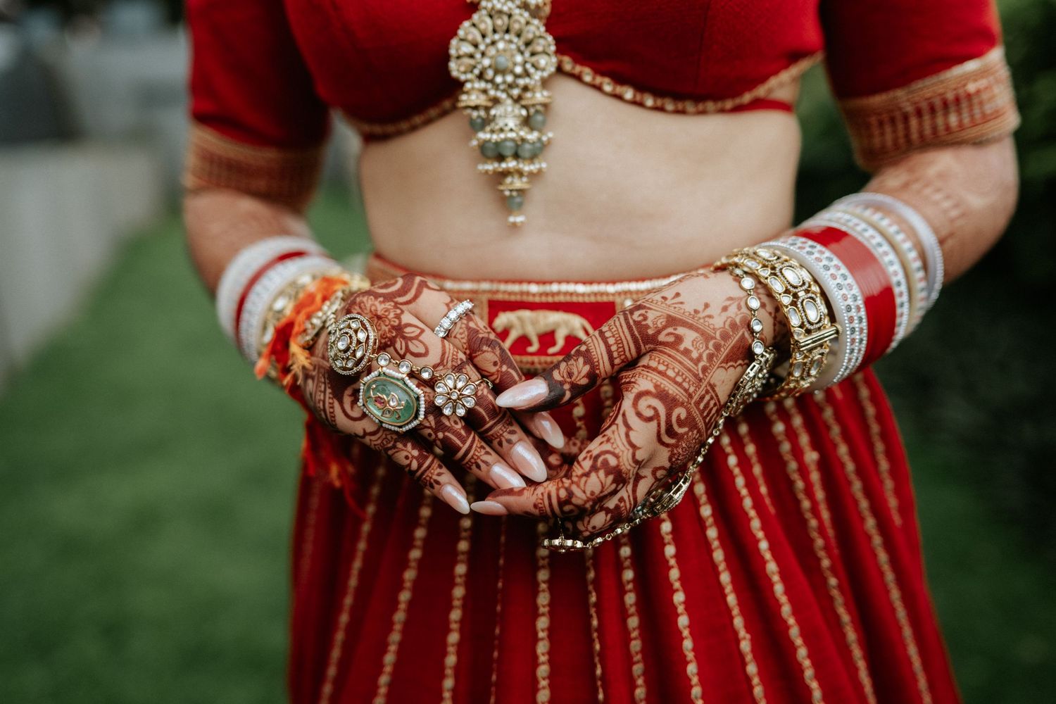 Indian bride showcasing intricate mehndi henna hands adorned with gold bangles and rings wearing red bridal lehenga.