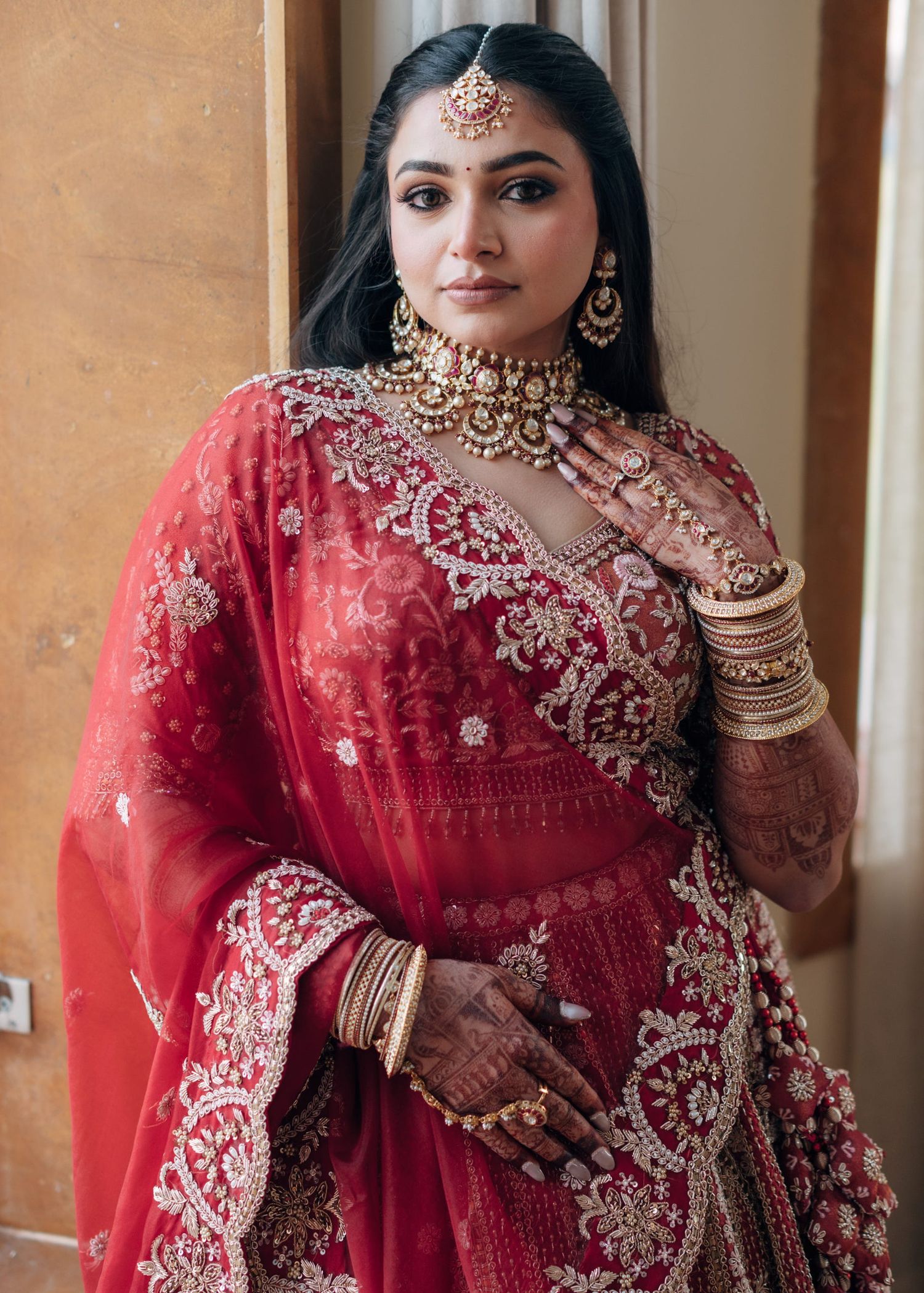 A bride in red lehenga with gold embroidery, wearing kundan jewelry, maang tikka, and mehndi on her hands.