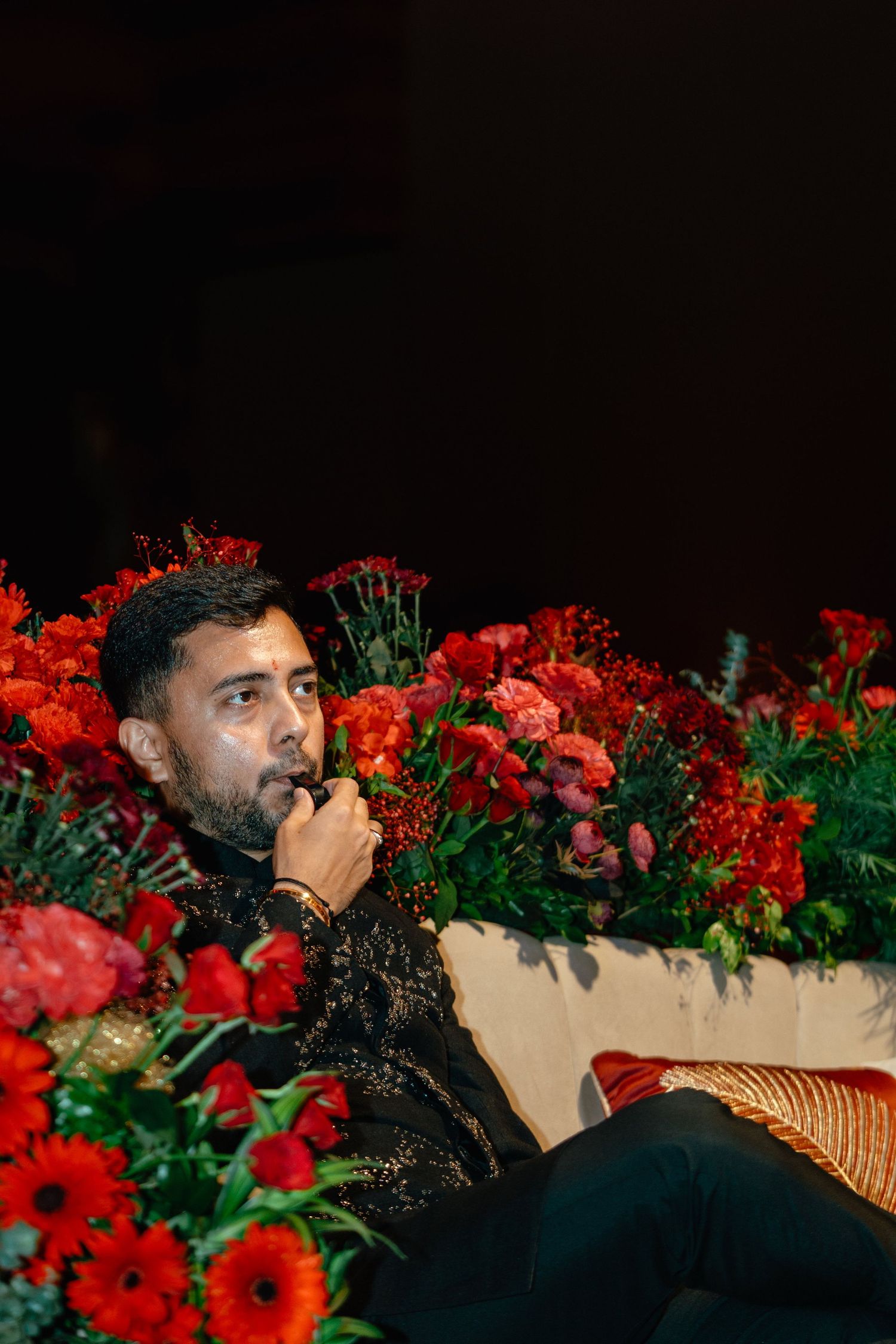 Man in black embroidered outfit seated among vibrant red floral arrangements at an elegant event.