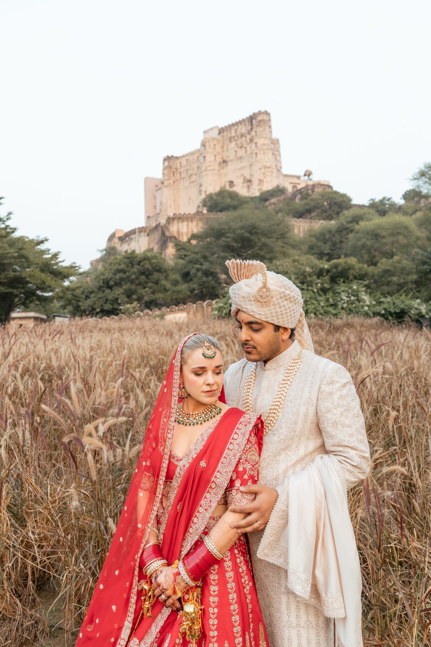 Couple in red and cream wedding attire pose in front of historic ruins and tall grass.
