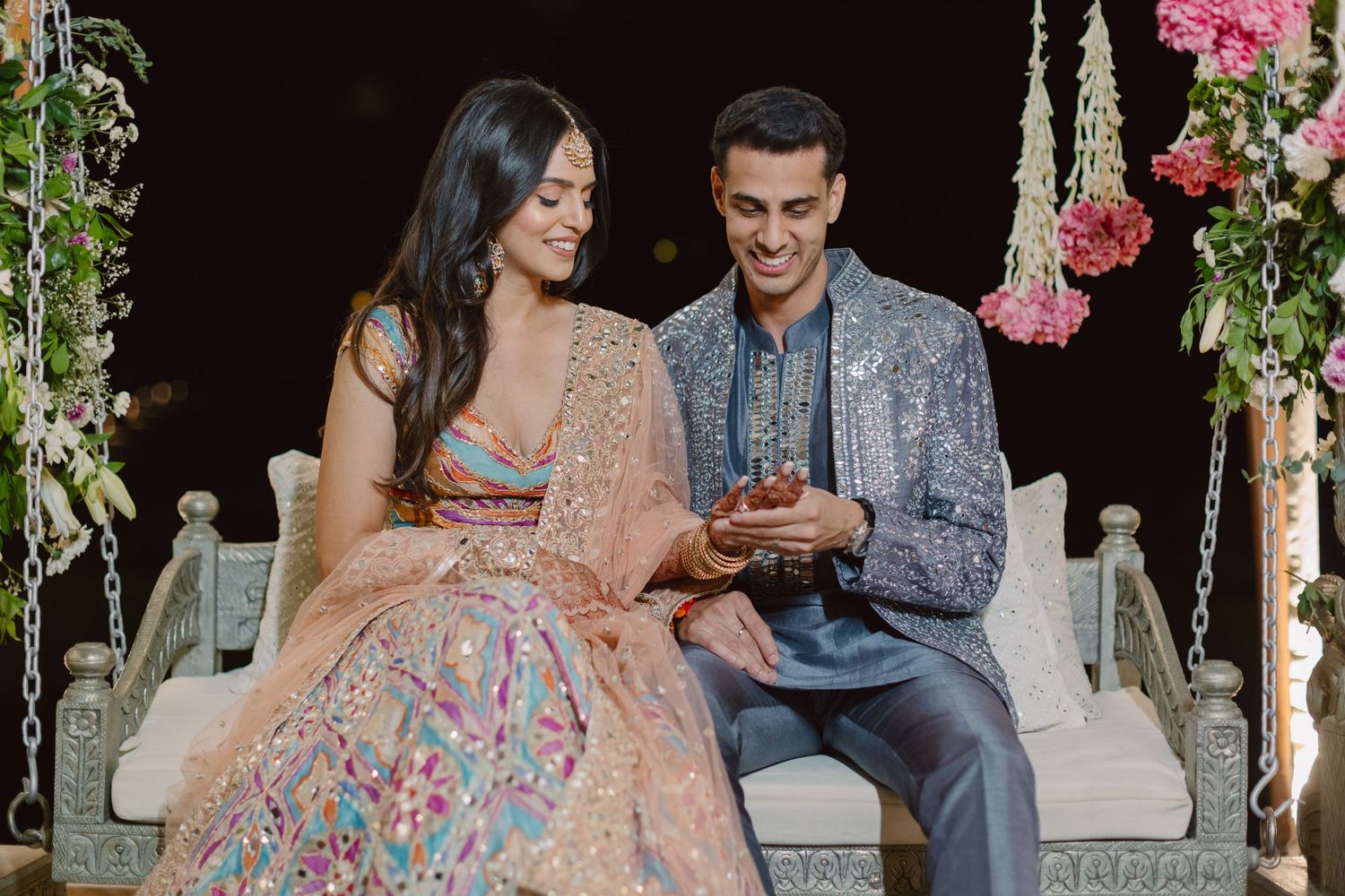 A couple in elegant Indian wedding attire shares a moment on a decorated swing with pink flowers.
