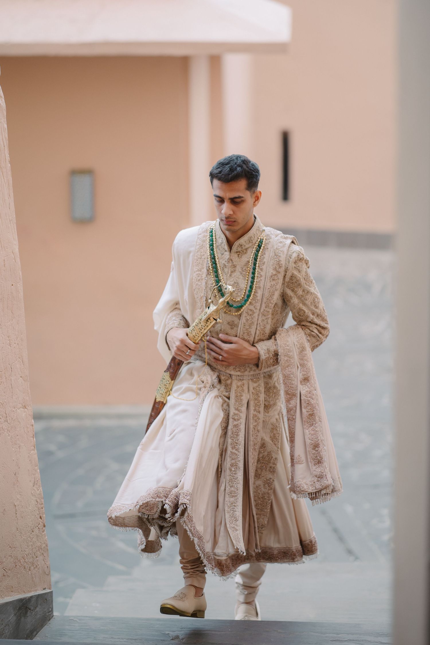 Groom in elegant ivory sherwani with green beaded necklace walking through courtyard.