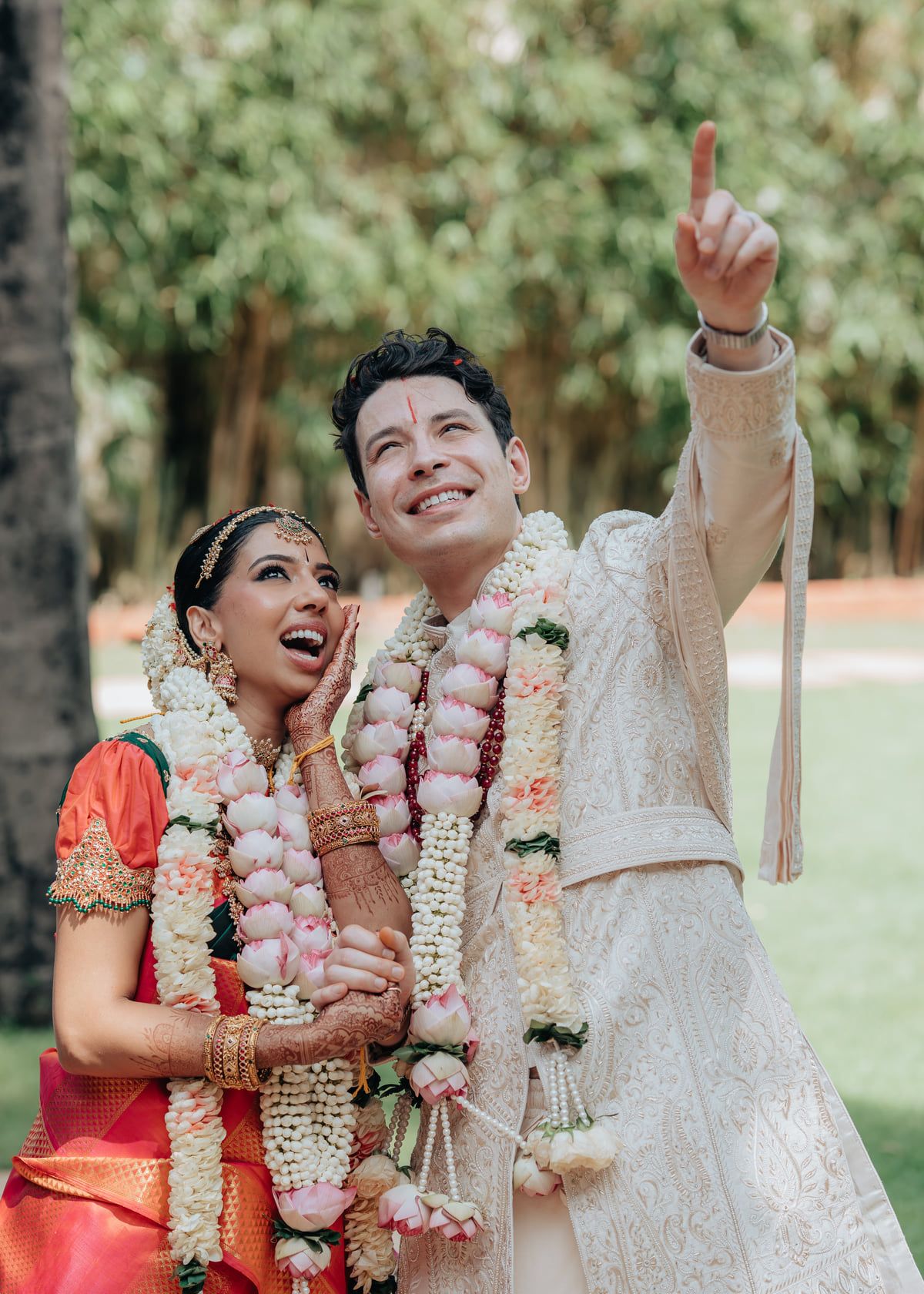 Joyful wedding couple wearing traditional garlands and ceremonial attire outdoors.