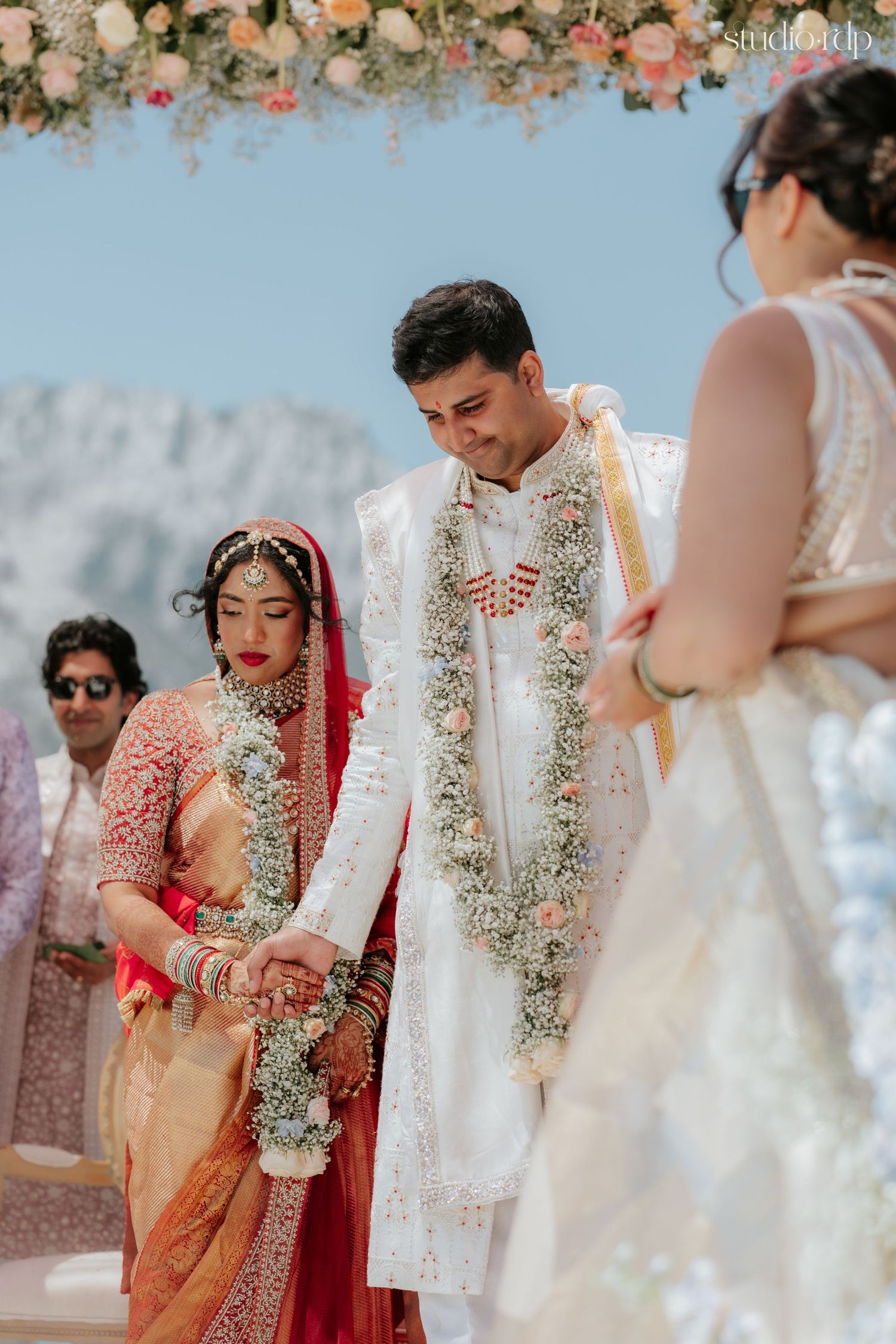 Indian bride in red saree and groom in white sherwani exchange garlands at traditional Hindu wedding ceremony.