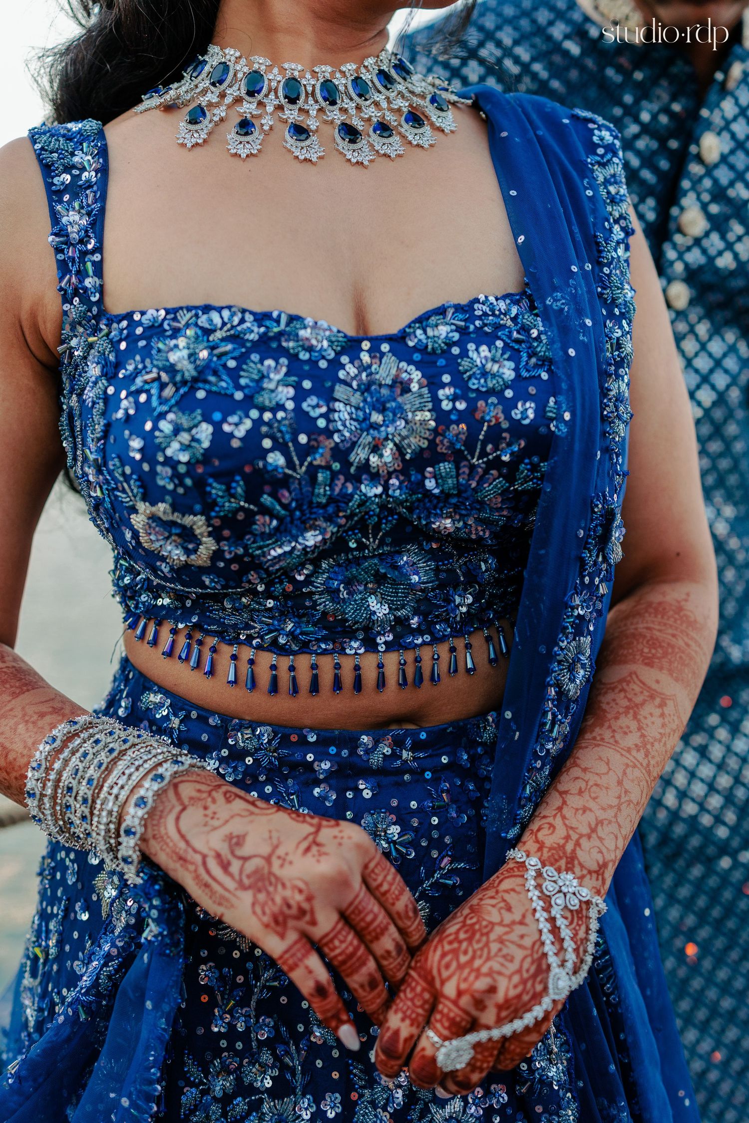Bride in royal blue embroidered lehenga with floral motifs, diamond necklace, bangles, and intricate mehndi on hands.