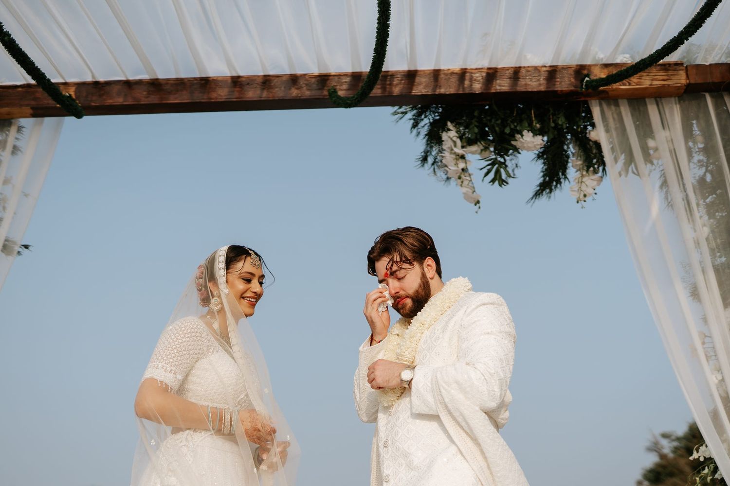 Bride and groom in white attire exchange wedding vows under a floral arch with blue sky backdrop.