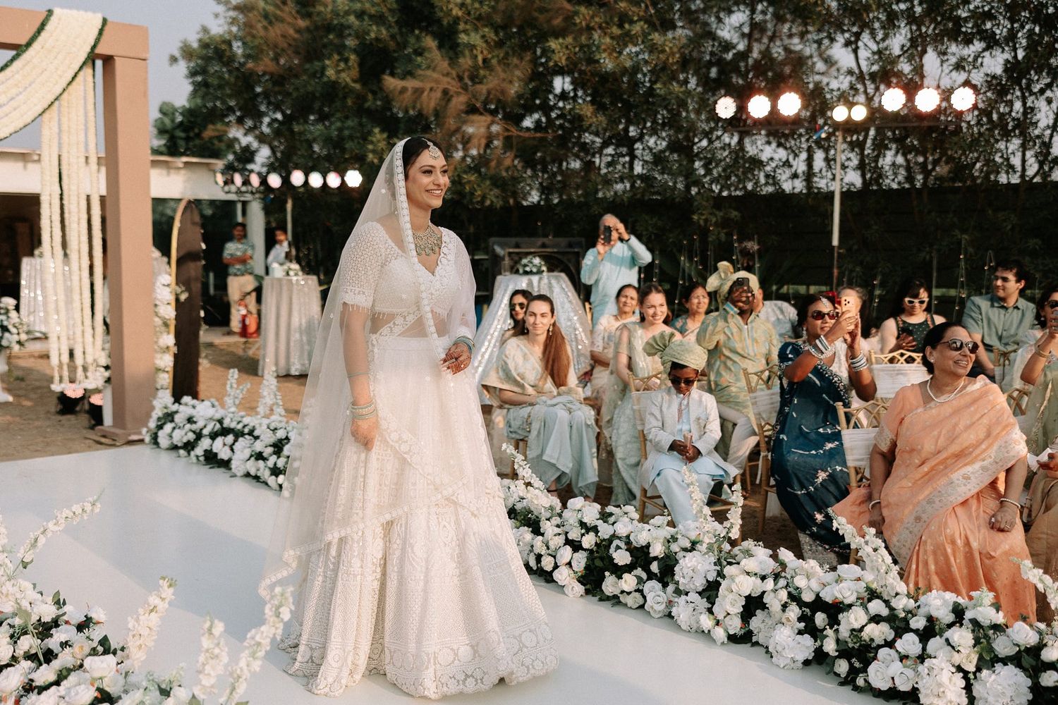 Bride in white lehenga and veil walking down floral aisle at outdoor wedding ceremony.