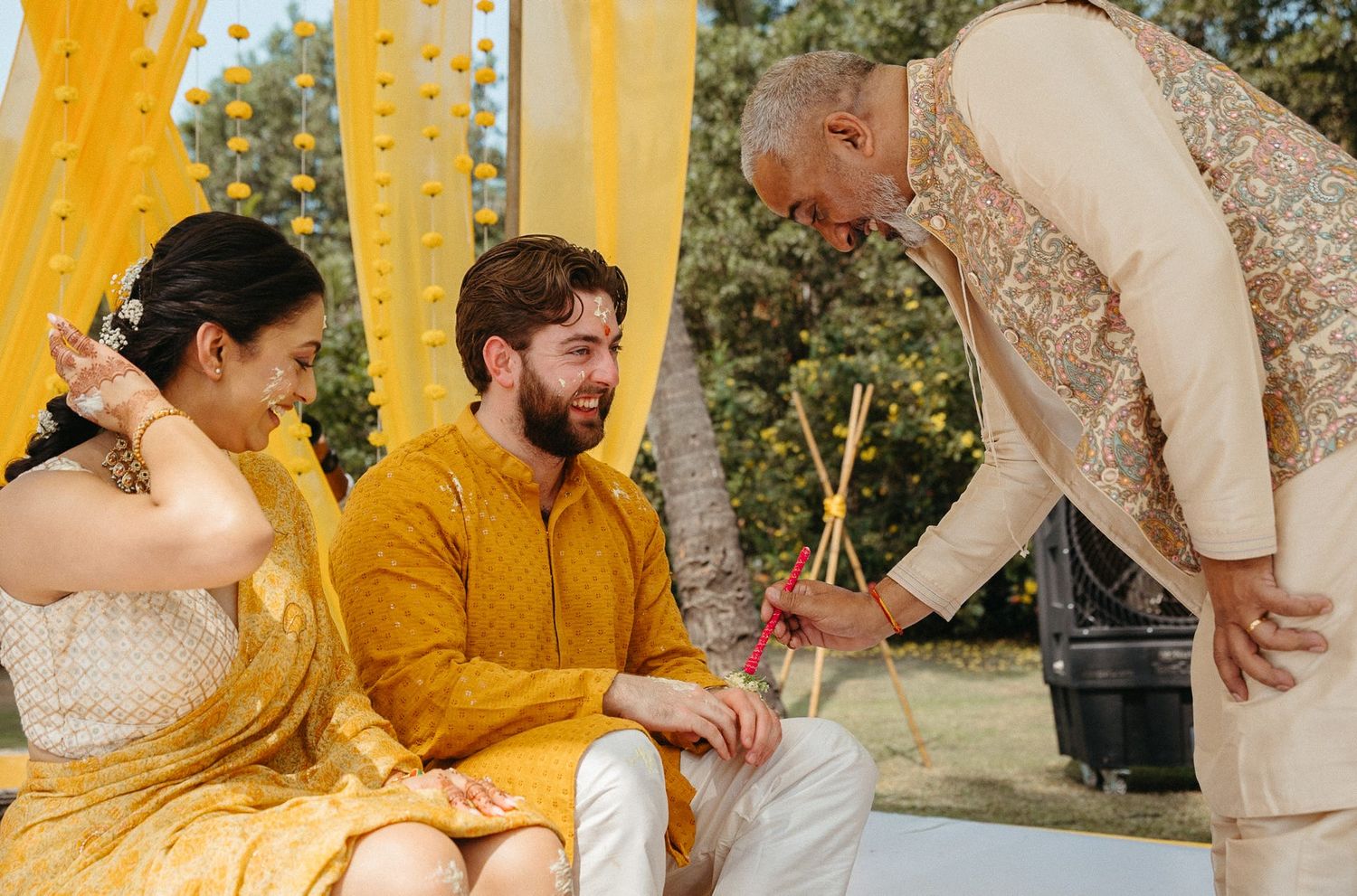 Couple in yellow outfits during Indian wedding Haldi ceremony with elder blessing them outdoors.