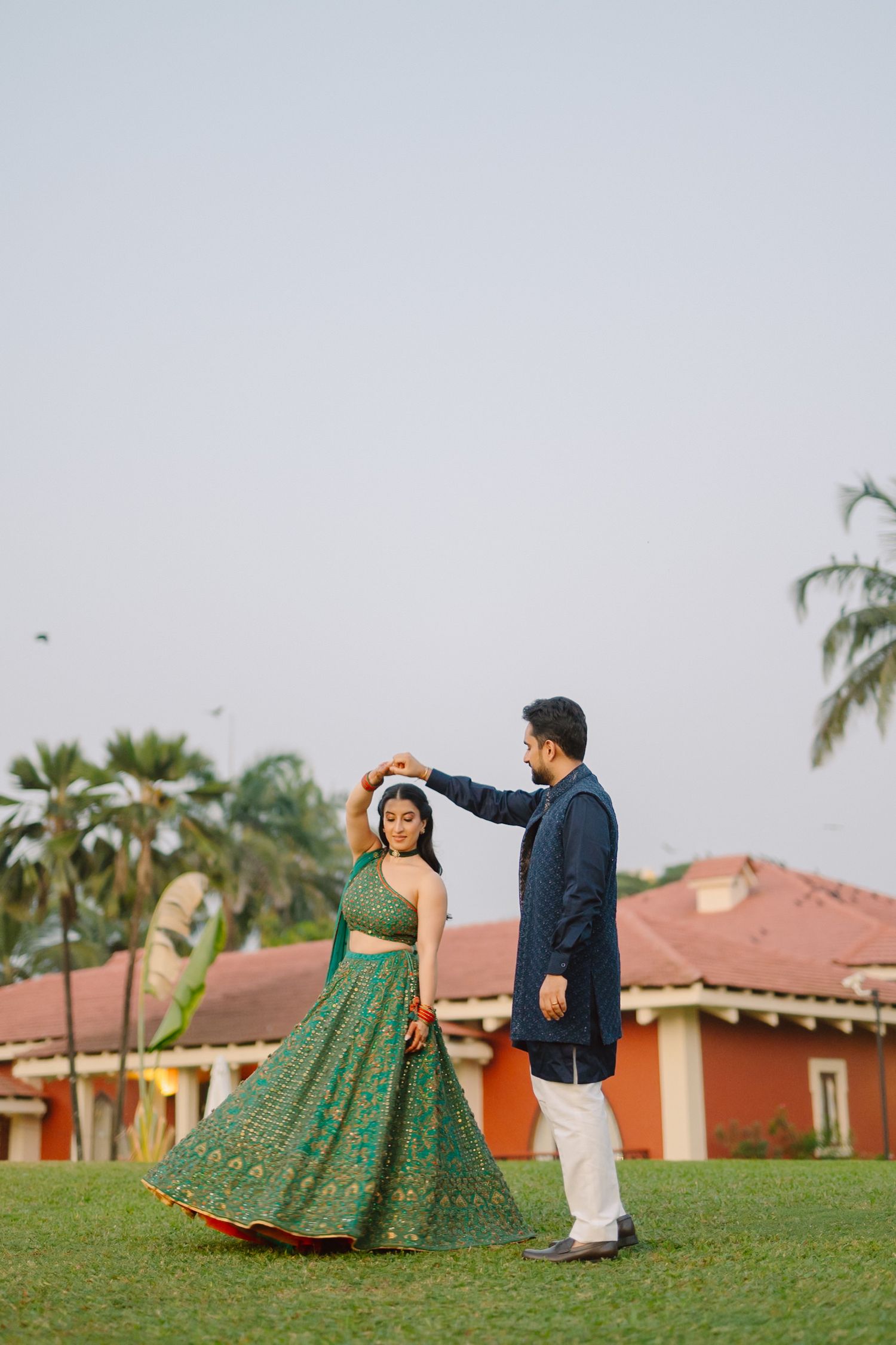 A couple dances together on a grassy lawn with palm trees and buildings in the background.