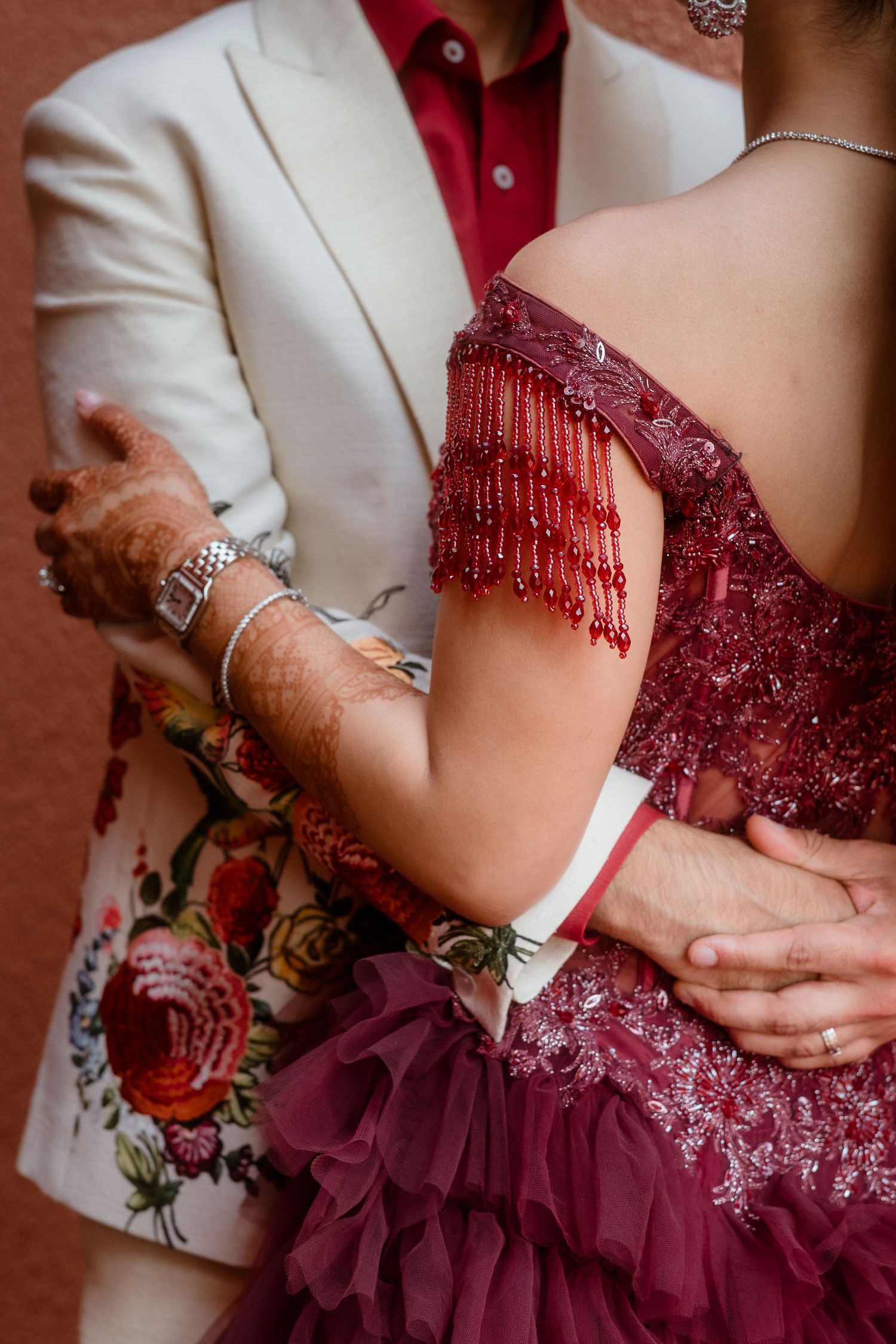 Close-up details of hands and a burgundy beaded wedding dress with white suit jacket.