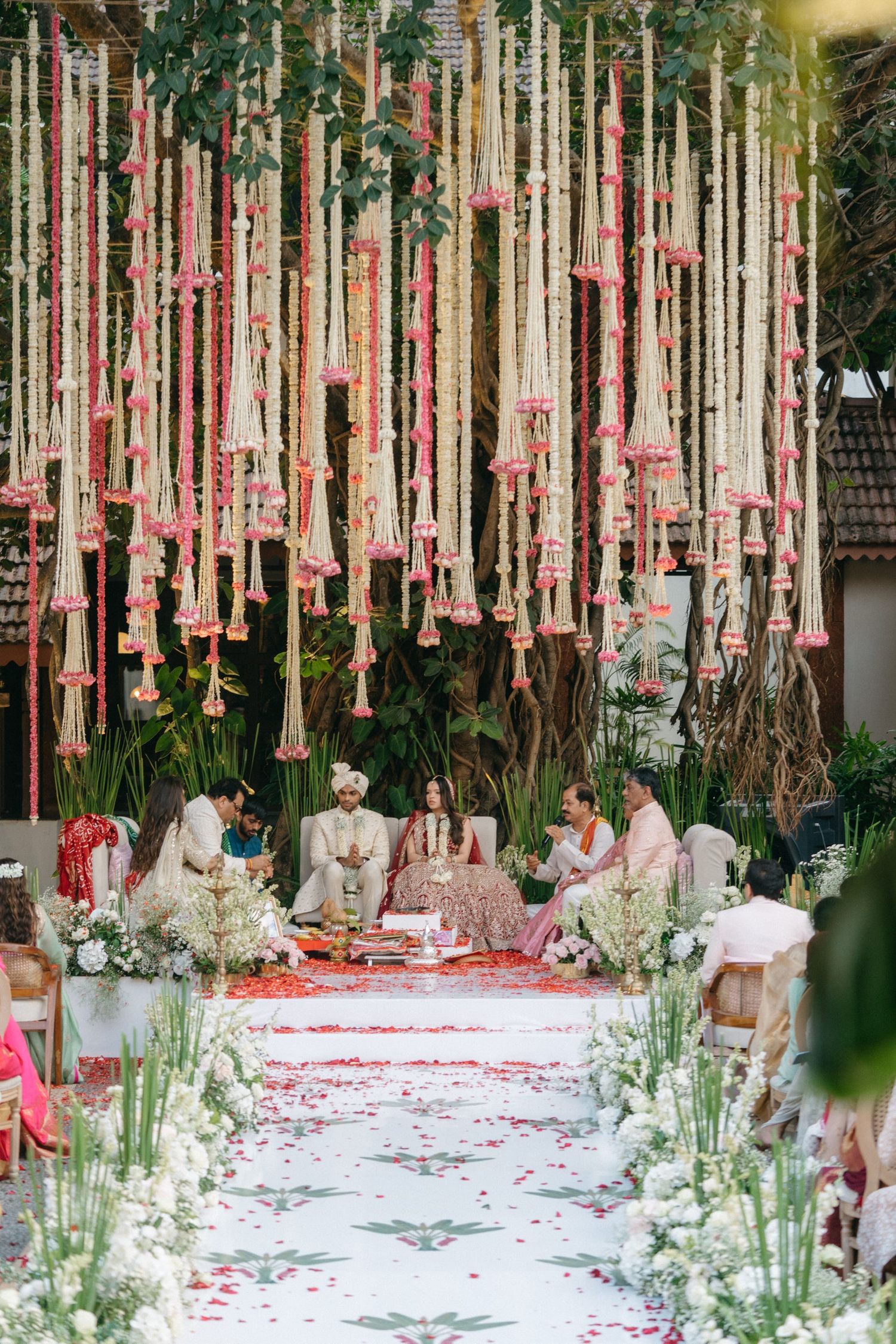 Indian wedding ceremony under floral canopy with hanging marigold garlands, bride and groom at mandap surrounded by white flowers.