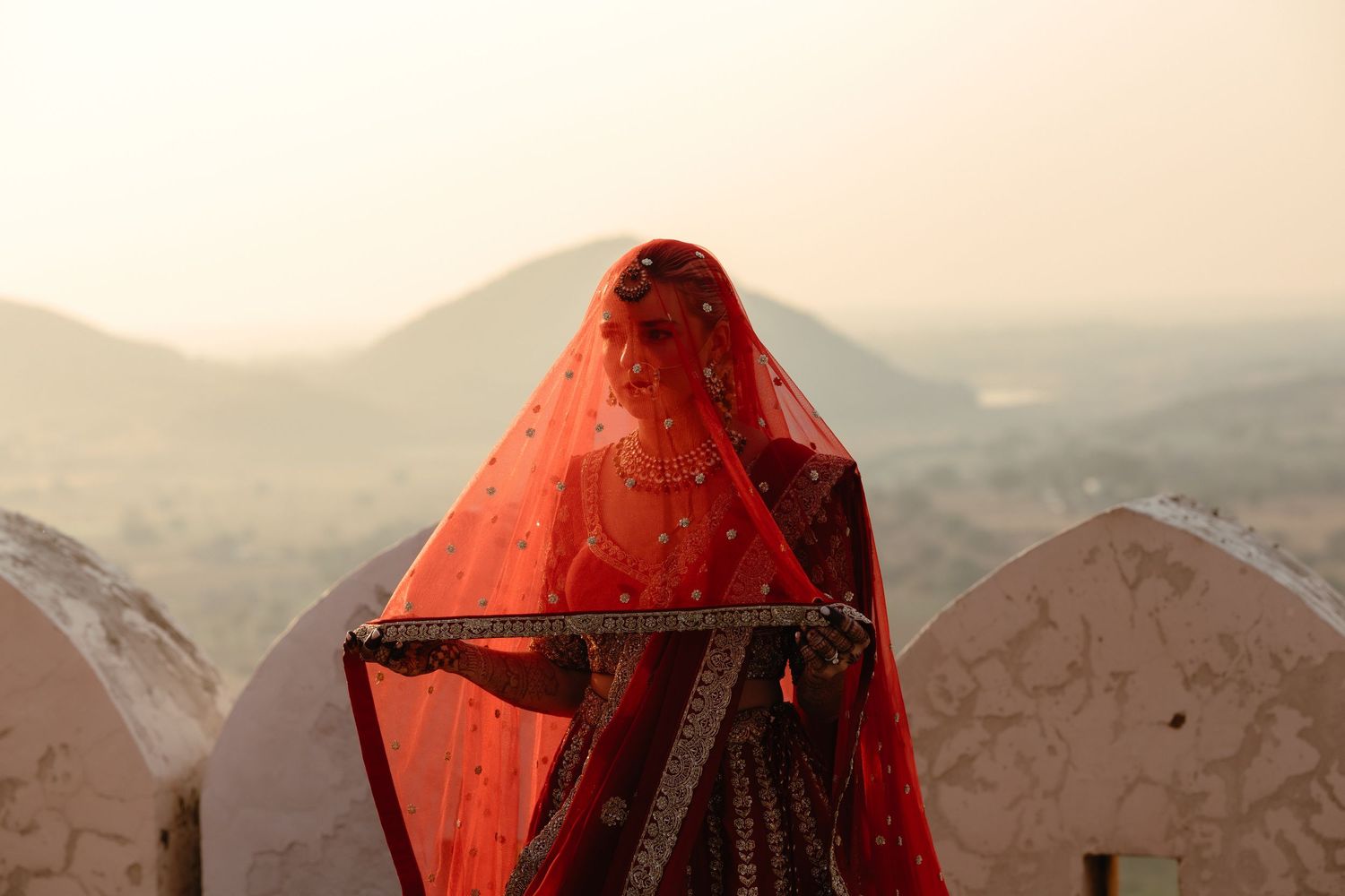 Woman in traditional red Indian bridal saree and veil standing on a terrace with misty mountain landscape.