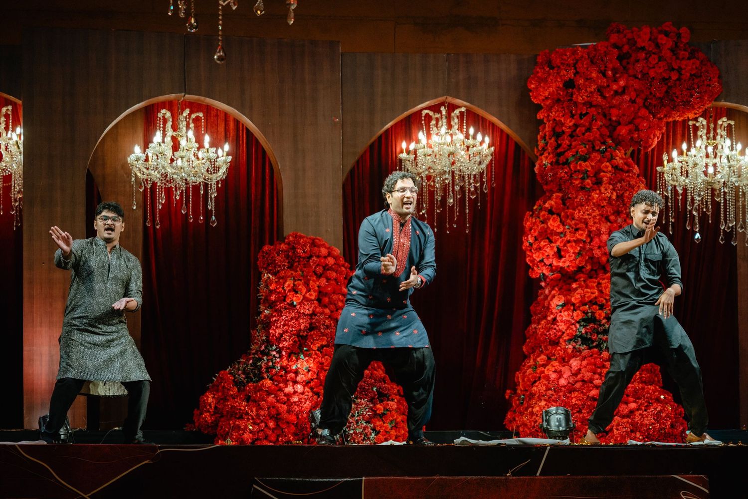Three performers on stage with red floral backdrop, chandeliers, and dramatic red curtains in a theatrical production.