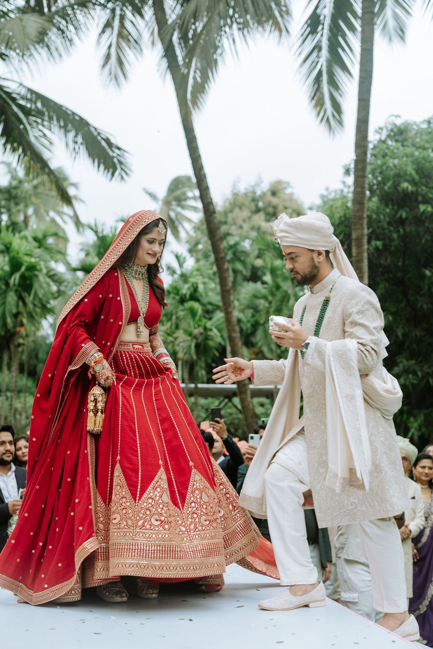 Bride in red lehenga and groom in ivory sherwani performing Hindu wedding rituals outdoors with palm trees.