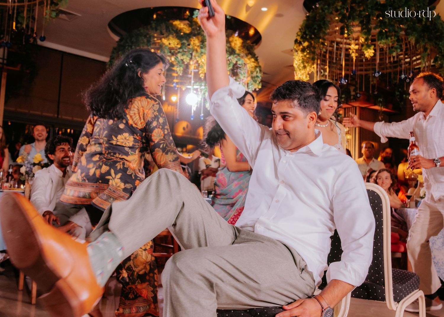 Joyful couple dancing at a wedding reception, man in white shirt raising hand, woman in floral outfit, festive venue.