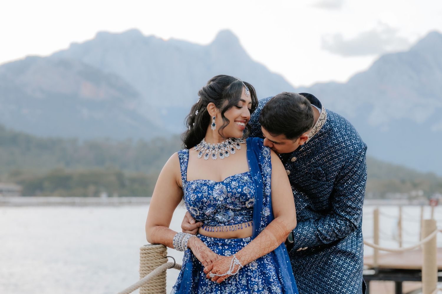 Indian couple in blue traditional attire posing on a dock with mountain lake backdrop.